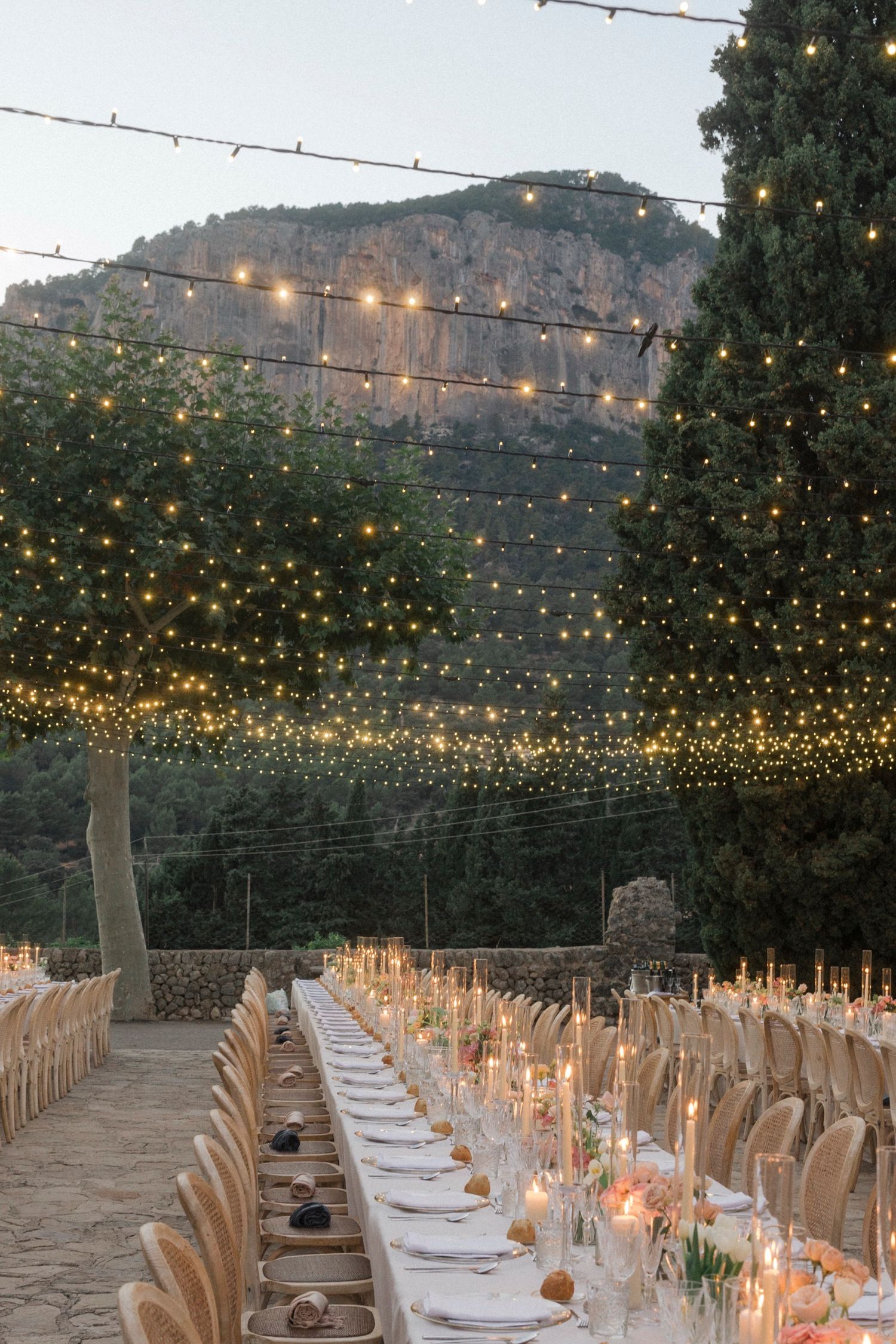 Elegant outdoor wedding reception setup with long tables under twinkling string lights at dusk in a mountain setting.