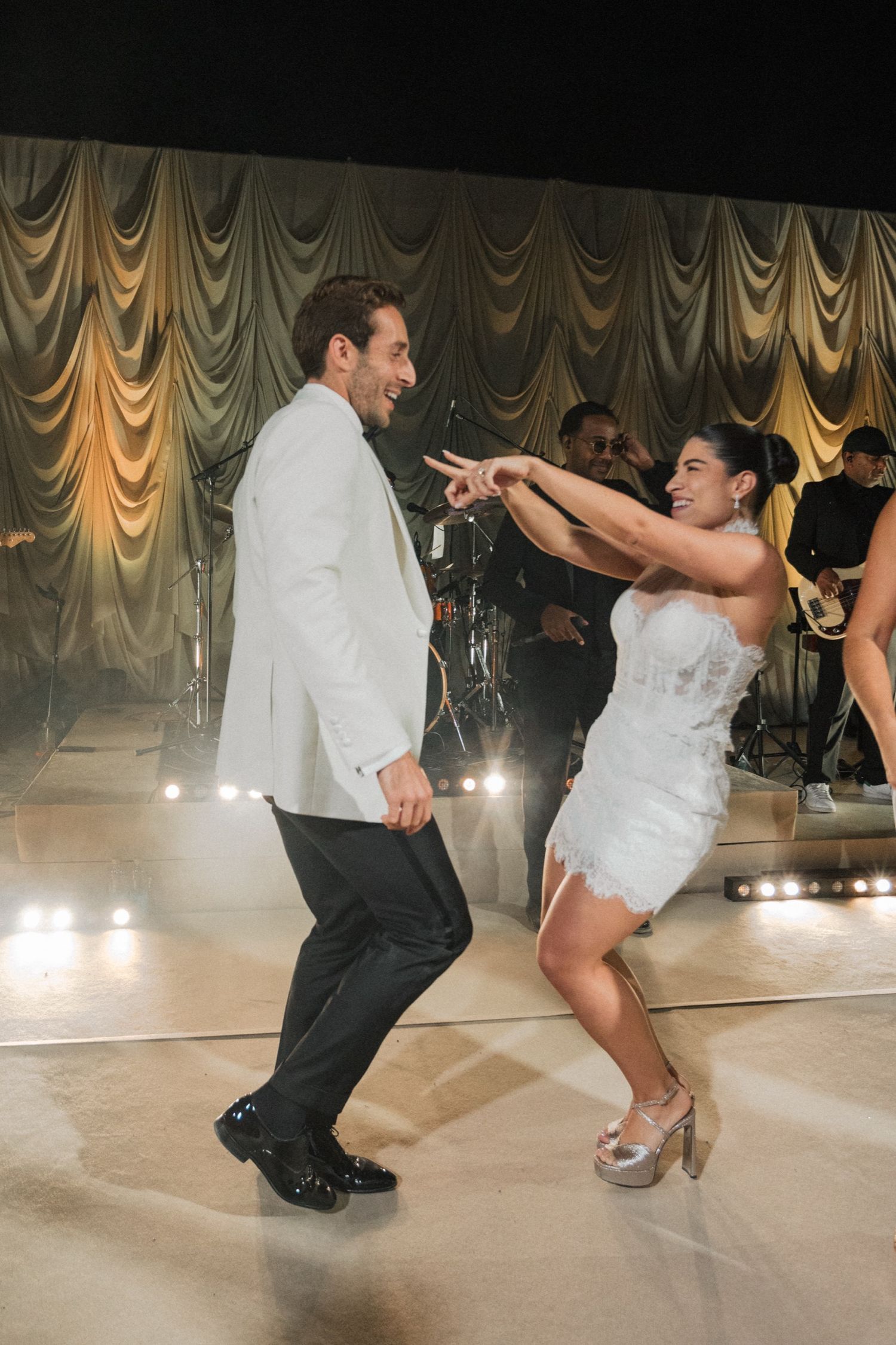 Couple performing energetic dance moves in formal attire on a ballroom floor with elegant draped curtains in background.