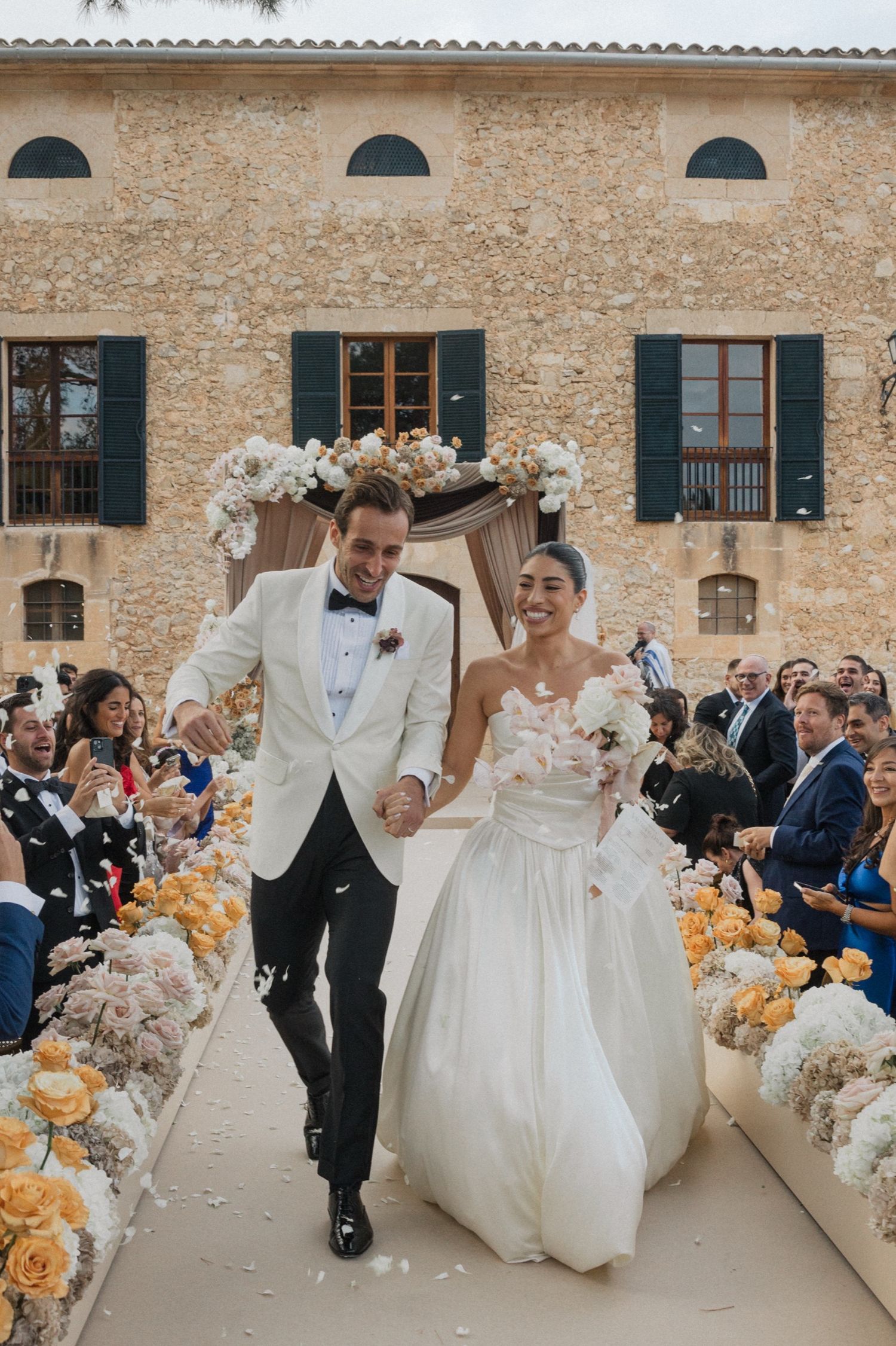 Joyful wedding party dancing and celebrating at an elegant stone venue with peach floral decorations.