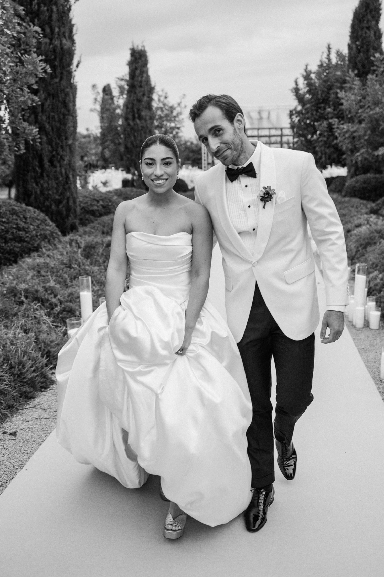 A bride in a strapless white gown and groom in white tuxedo jacket walk together on a garden path in black and white.