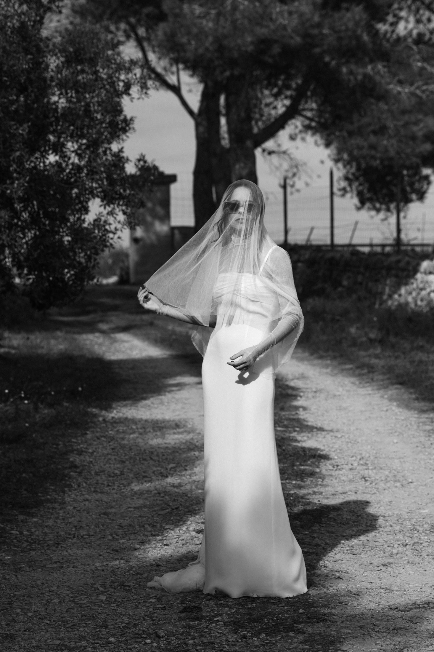 Black and white photo of a bride in a flowing white dress and veil standing on a garden path surrounded by trees.
