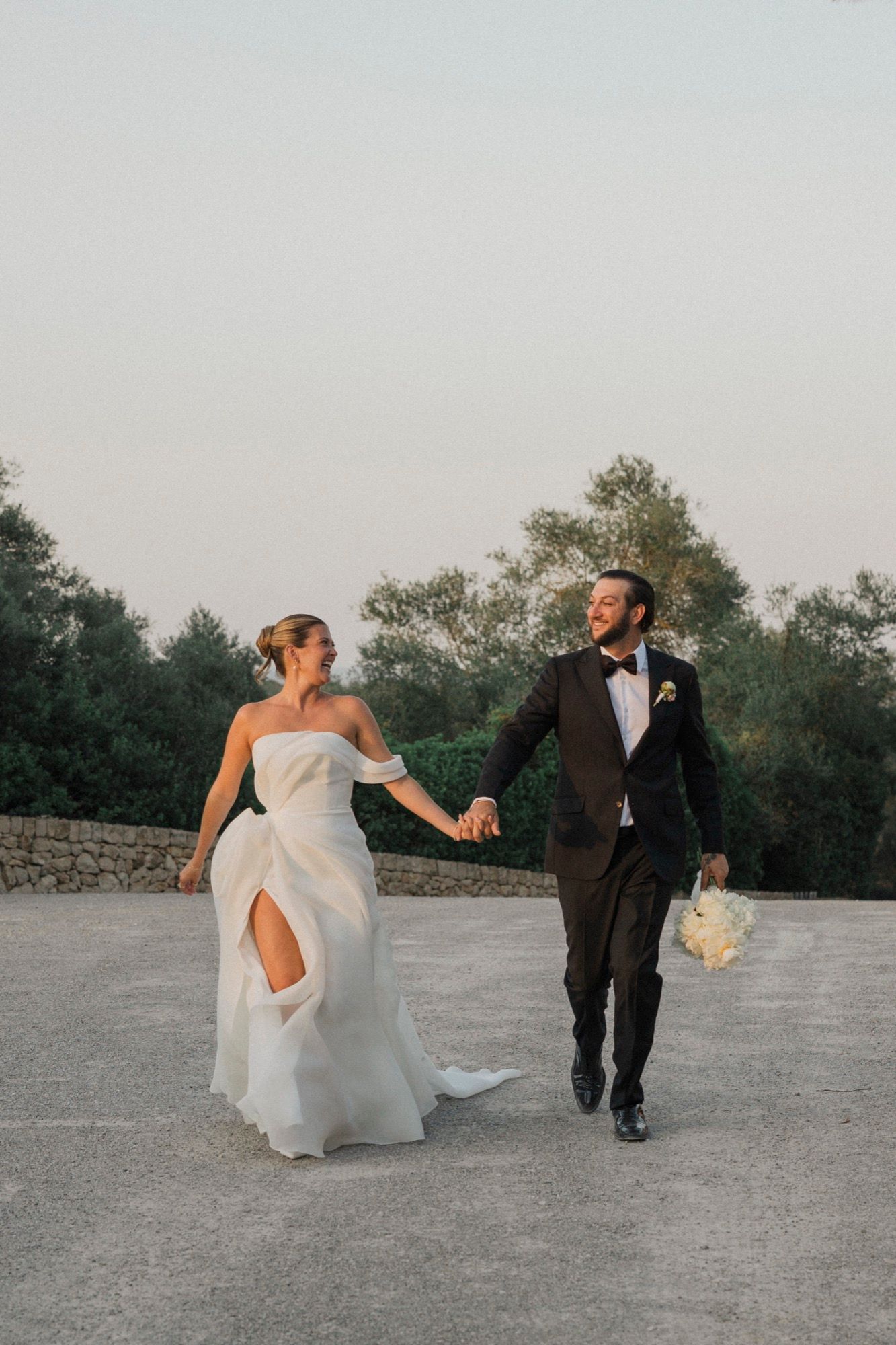 Bride in flowing white dress and groom in black tux walk together on outdoor stone terrace.