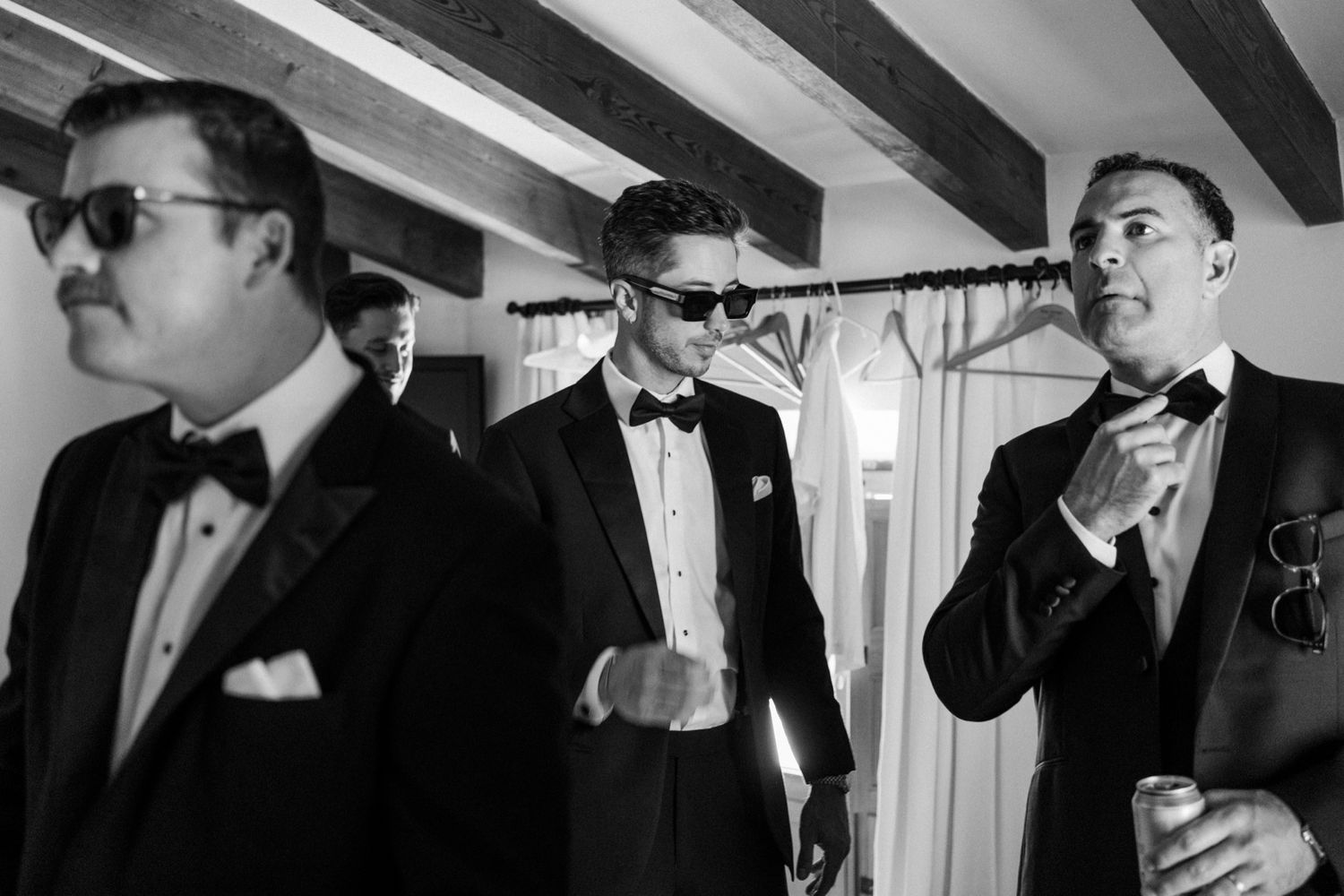 Groomsmen in black tuxedos and sunglasses pose together in a stylish black and white photo.