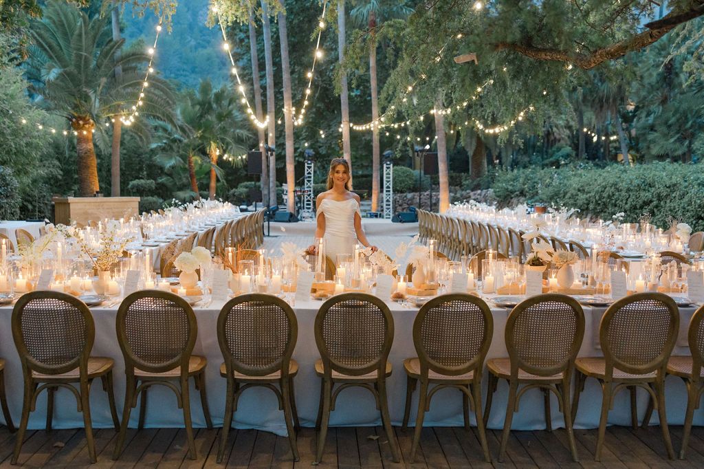 Rustic wooden chairs line a long outdoor wedding reception table under string lights and trees.