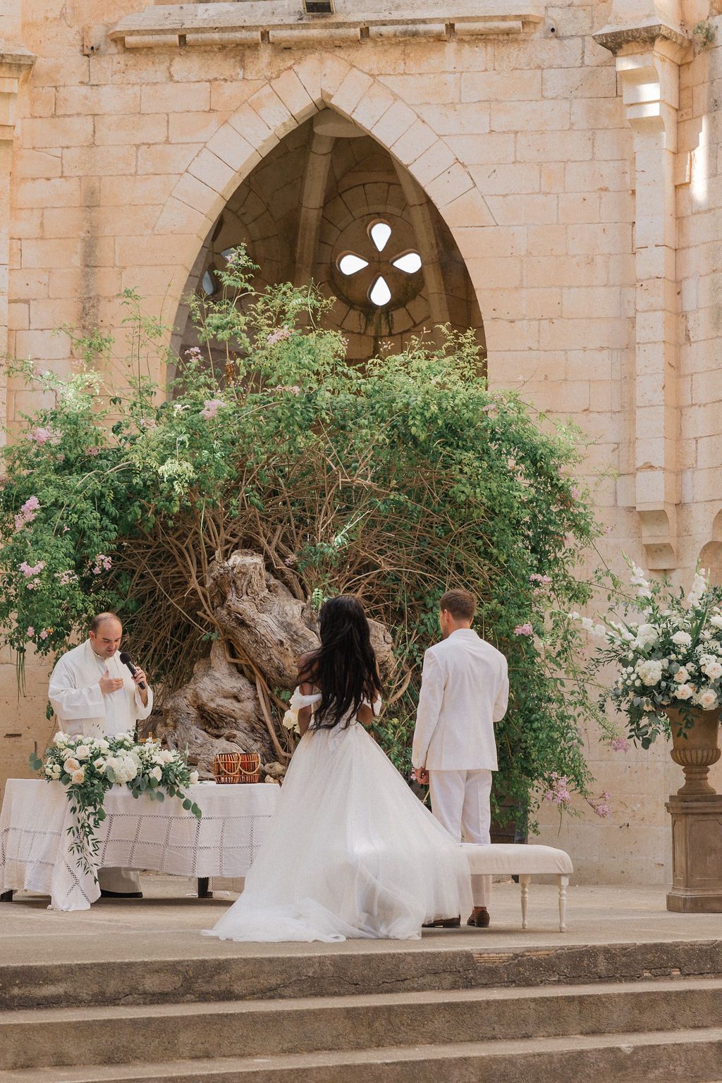 Wedding ceremony setup in a historic stone courtyard with floral decorations and white chairs.