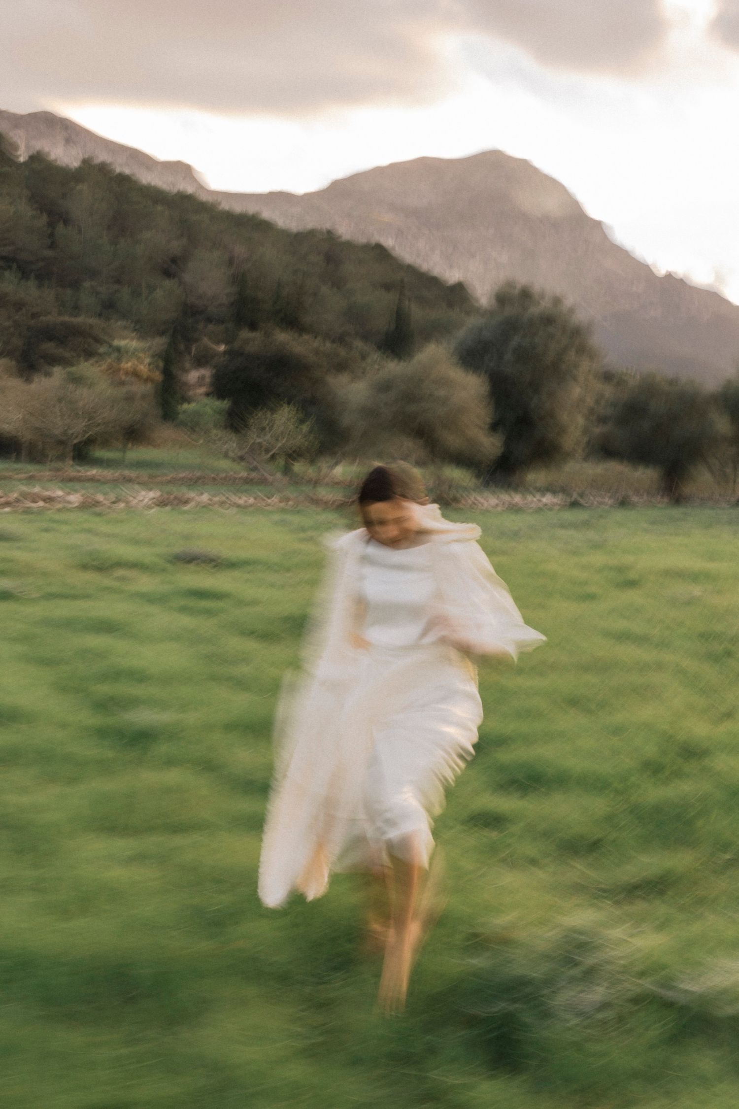 A figure in a flowing white dress leaps through a green meadow with misty mountains in the background.
