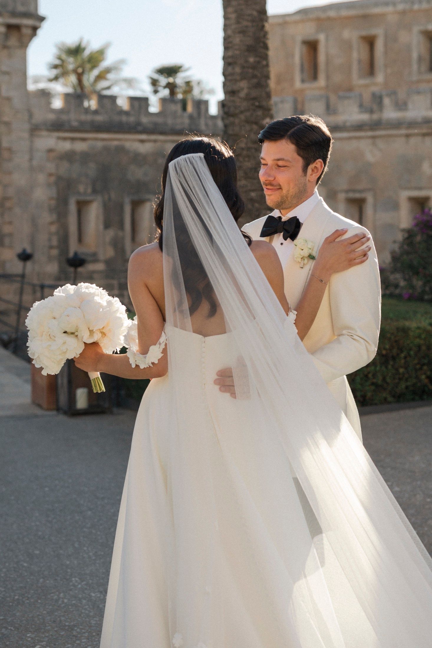 Bride and groom embrace outdoors at a castle venue, bride holding white bouquet with veil, groom in white tuxedo with bow tie.