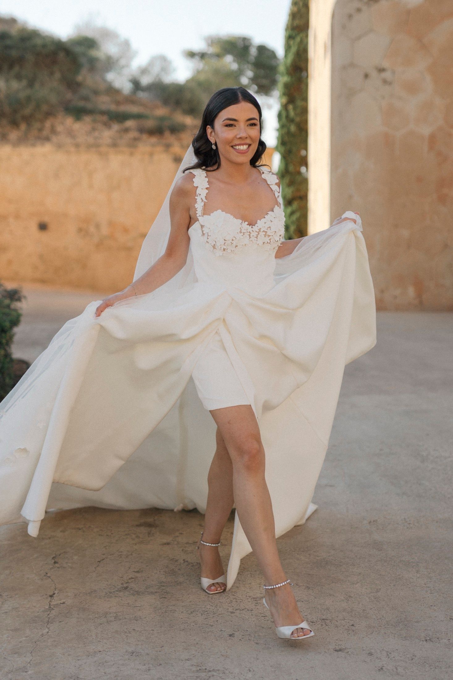 Smiling bride in short white lace wedding dress with flowing veil, posing outdoors in golden sunlight wearing strappy heels.