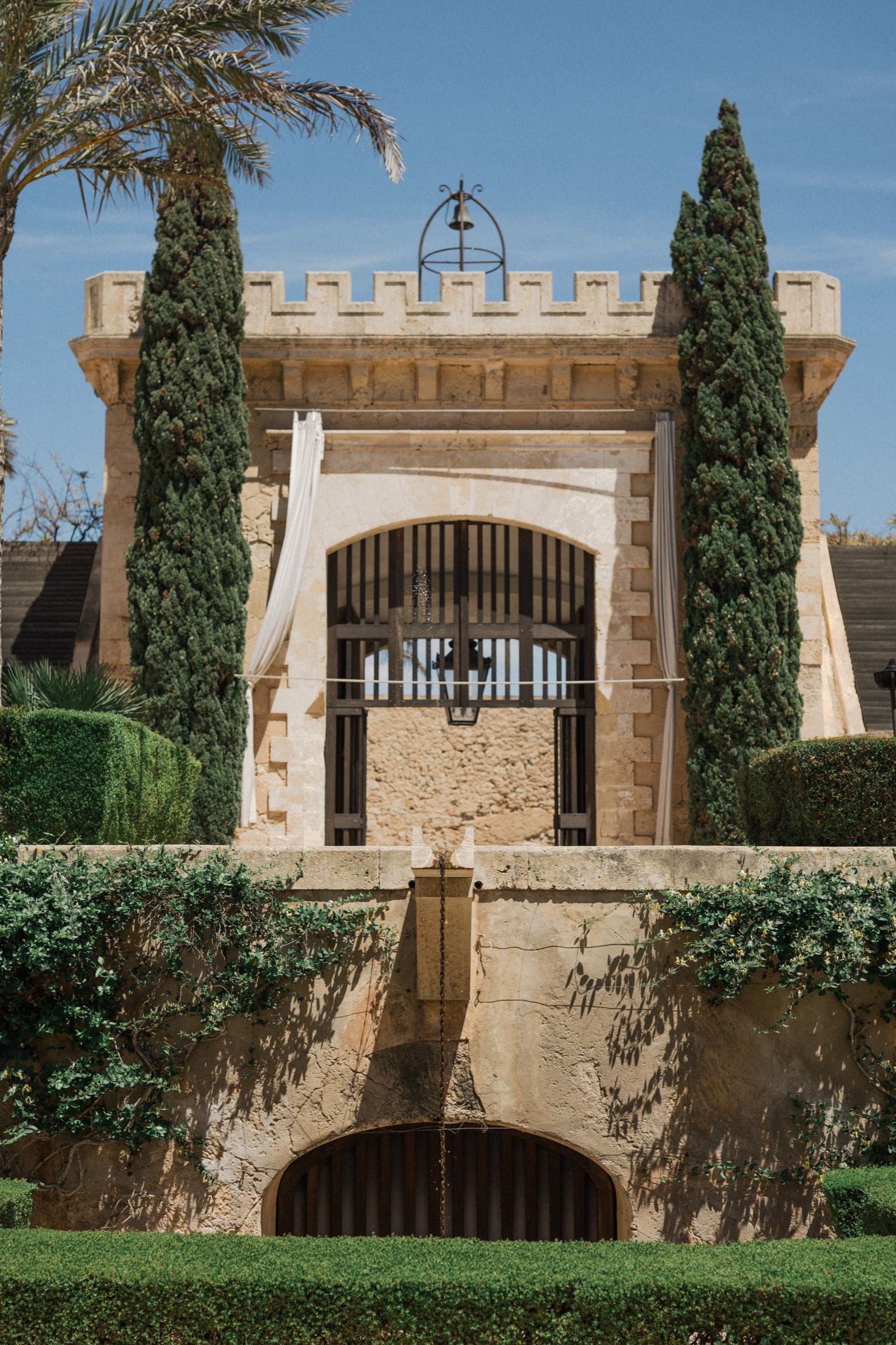 Historic stone castle gate with arched entrance, battlements, cypress trees, and lush green gardens under a blue sky.