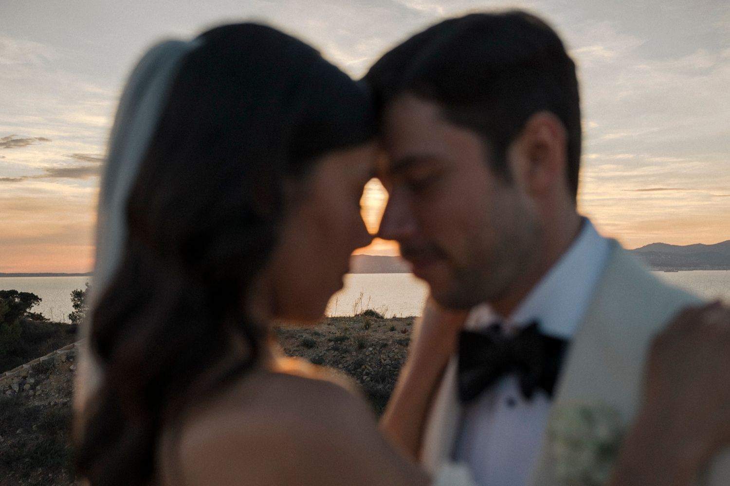 Bride and groom share an intimate moment at sunset by the sea, with warm golden light glowing between them.