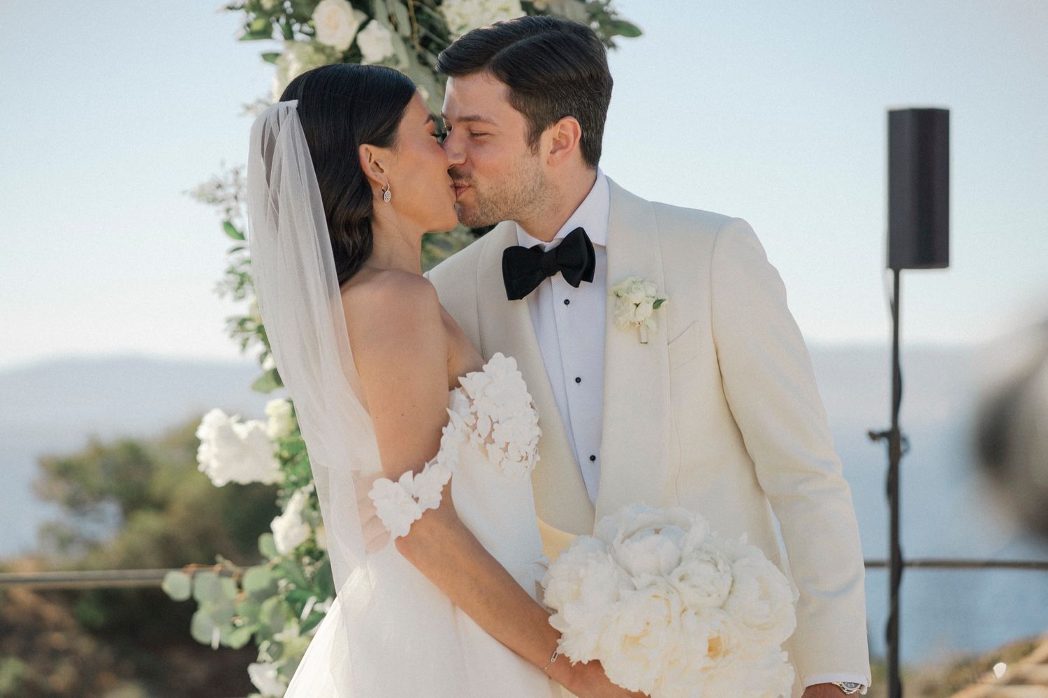 Bride and groom share a kiss at outdoor wedding ceremony, bride in white gown with veil, groom in ivory tuxedo with bow tie.