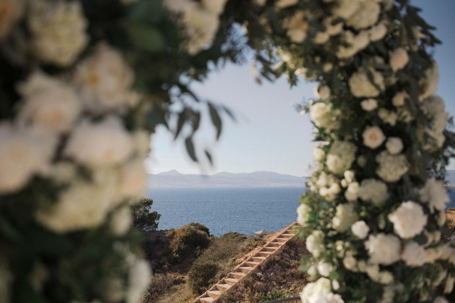Floral arch with white flowers frames a scenic ocean view at an outdoor wedding ceremony.