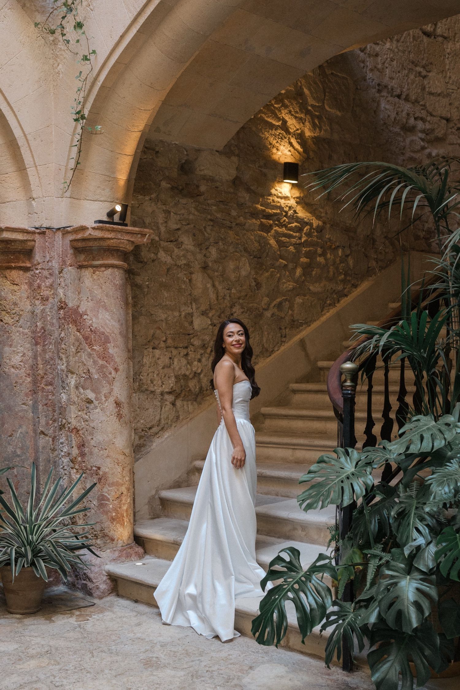A white wedding dress flows elegantly against stone archways and tropical plants in a rustic Mediterranean setting.