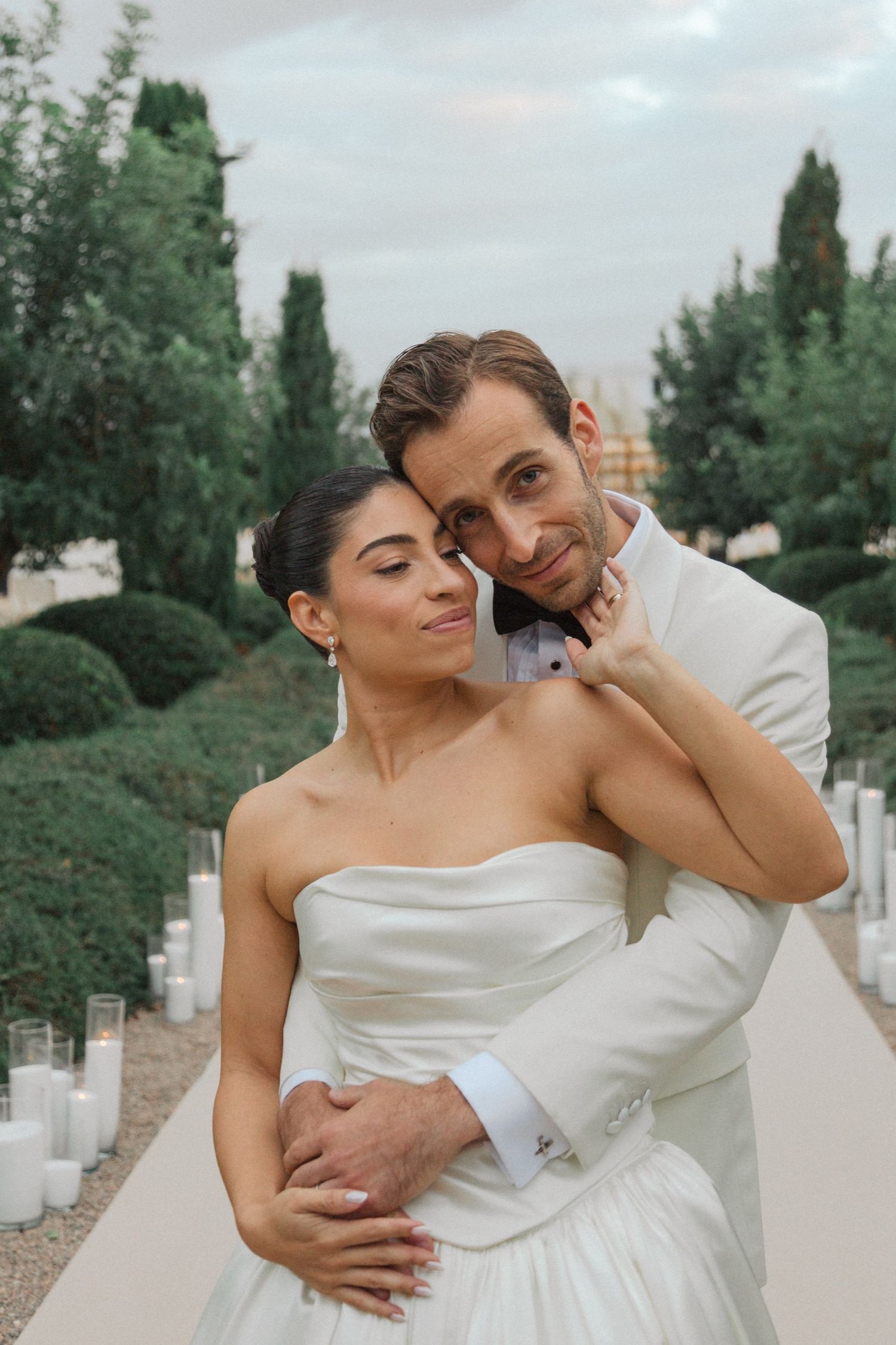 A romantic wedding couple embraces on a garden path lined with candles at dusk.