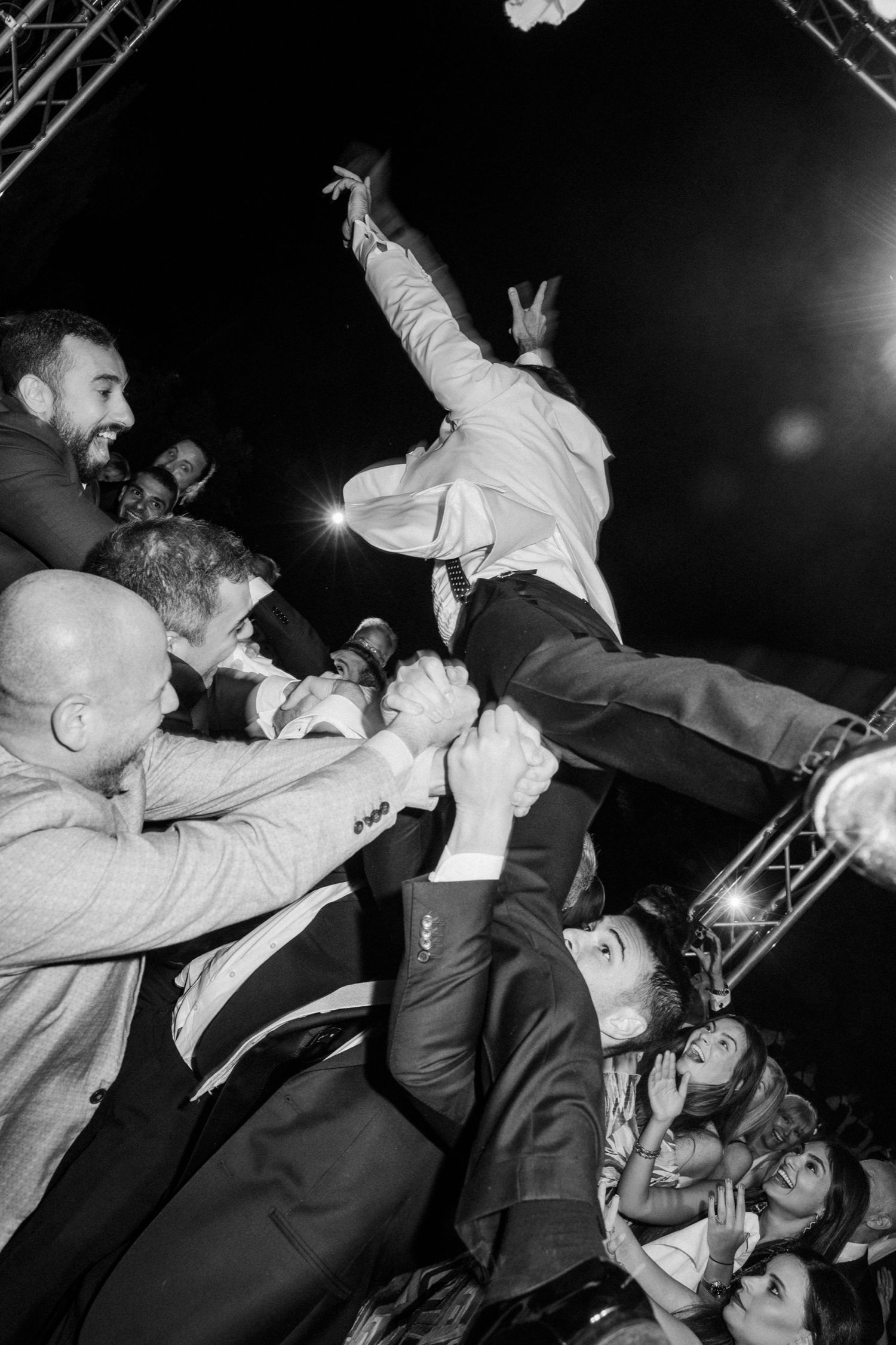 A crowd surfing performer leaps into the audience at a concert while fans reach up to catch them in black and white.