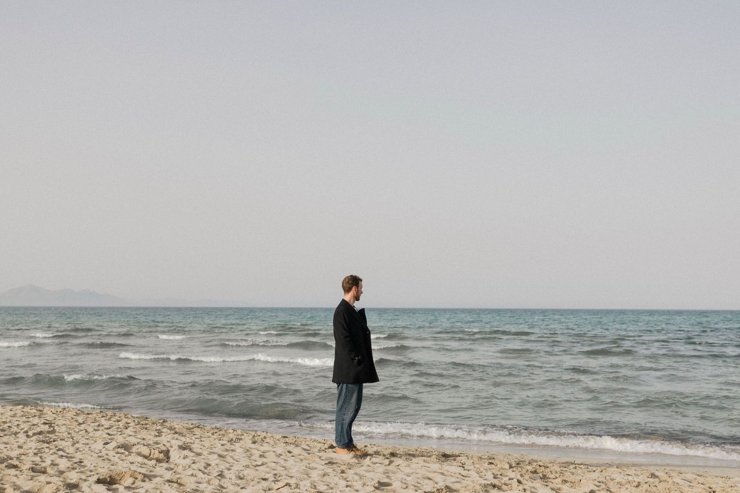 A minimalist beach scene with a solitary figure walking along the shoreline against an overcast sky.