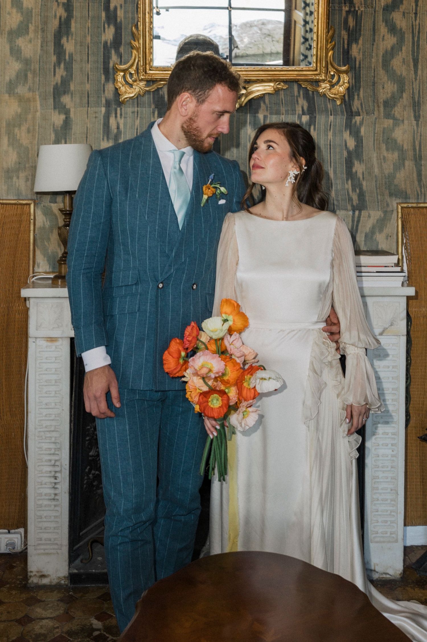 Bride in elegant white dress holds orange bouquet while gazing lovingly at groom in blue plaid suit at indoor venue.