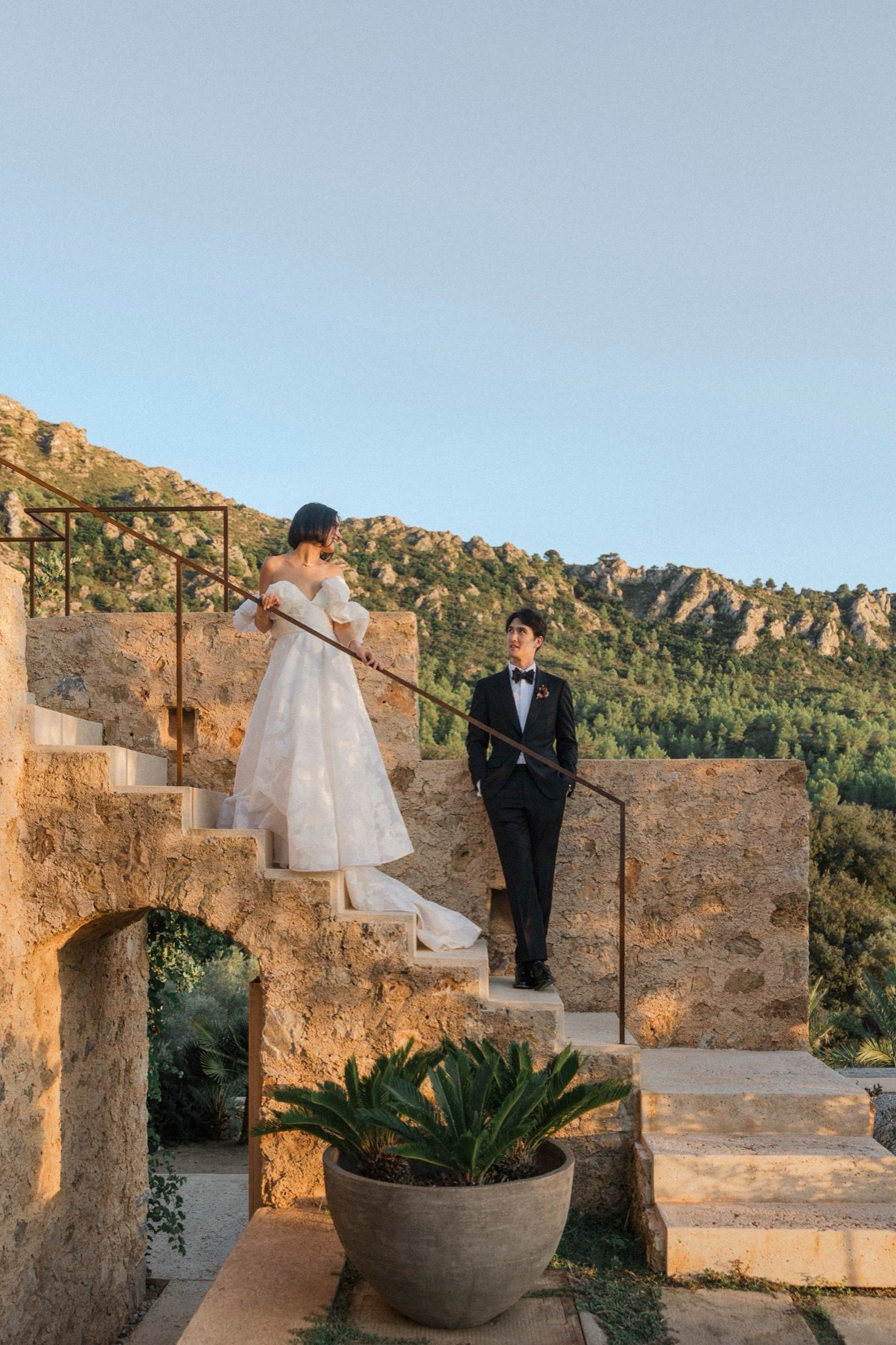 A wedding couple poses on rustic stone steps at a scenic outdoor venue during golden hour.