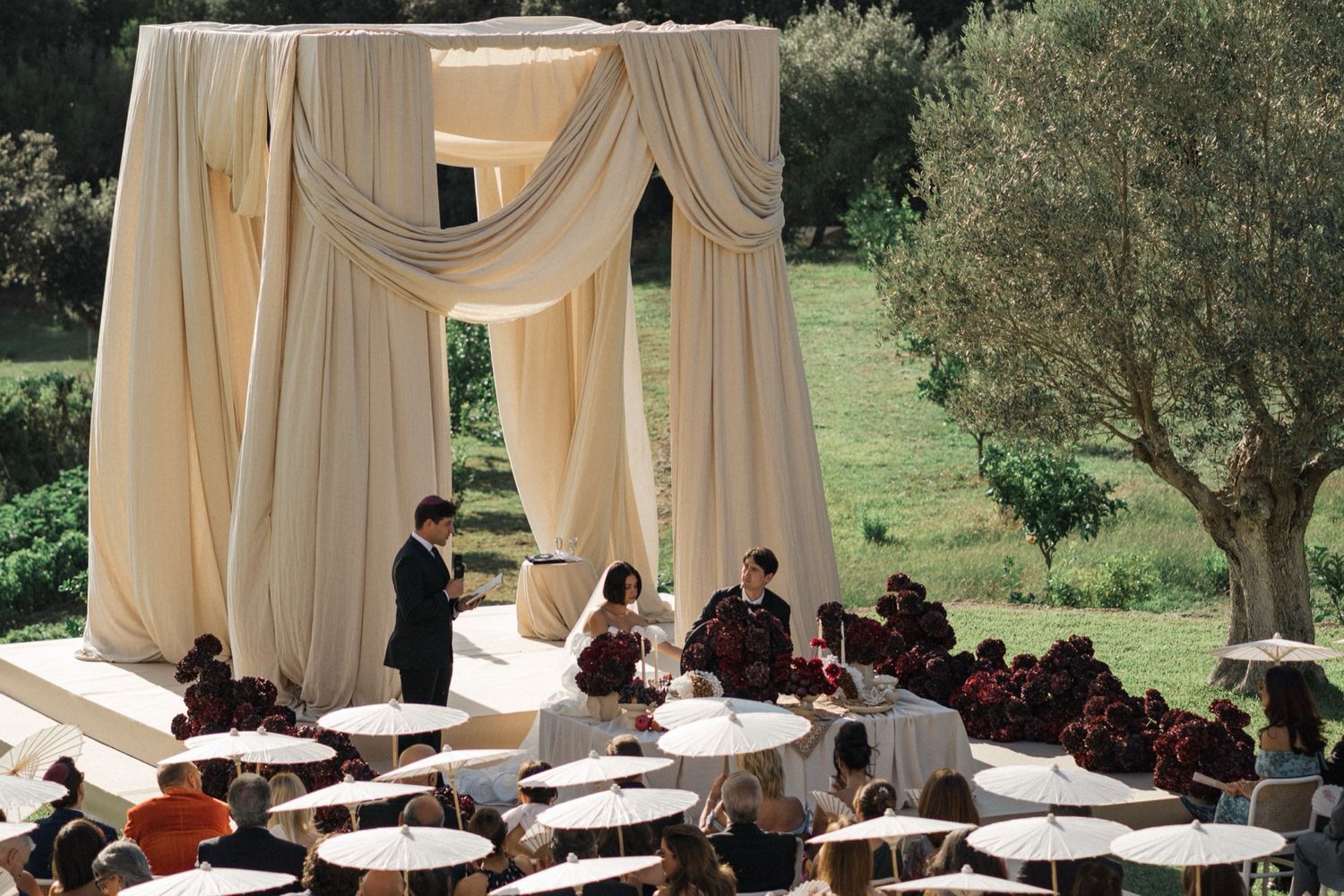 An elegant outdoor wedding ceremony setup with cream-colored drapes and white chairs under an olive tree.