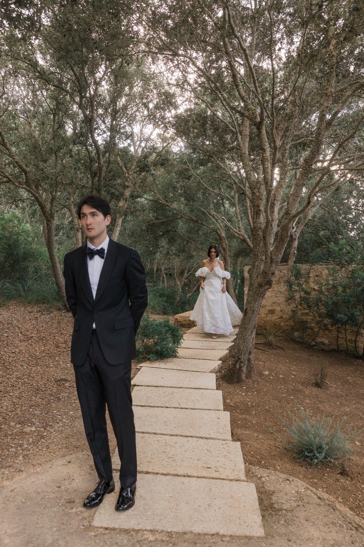A couple in formal wedding attire poses playfully on concrete steps surrounded by trees in an outdoor setting.