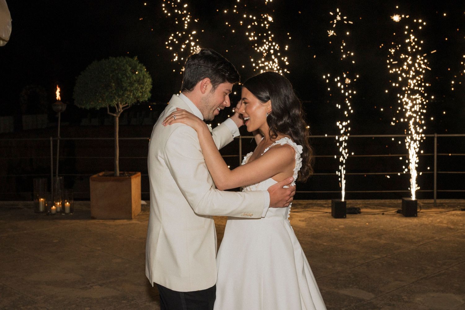Bride and groom share first dance with sparkling fireworks backdrop at elegant outdoor night wedding celebration.