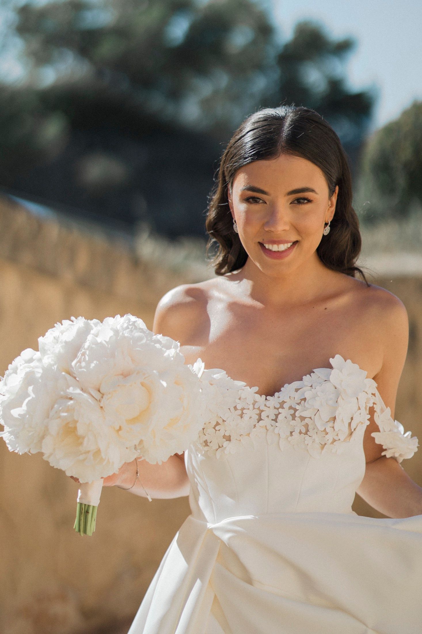 Smiling bride in off-shoulder lace wedding gown holding white peony bouquet outdoors in natural sunlight.