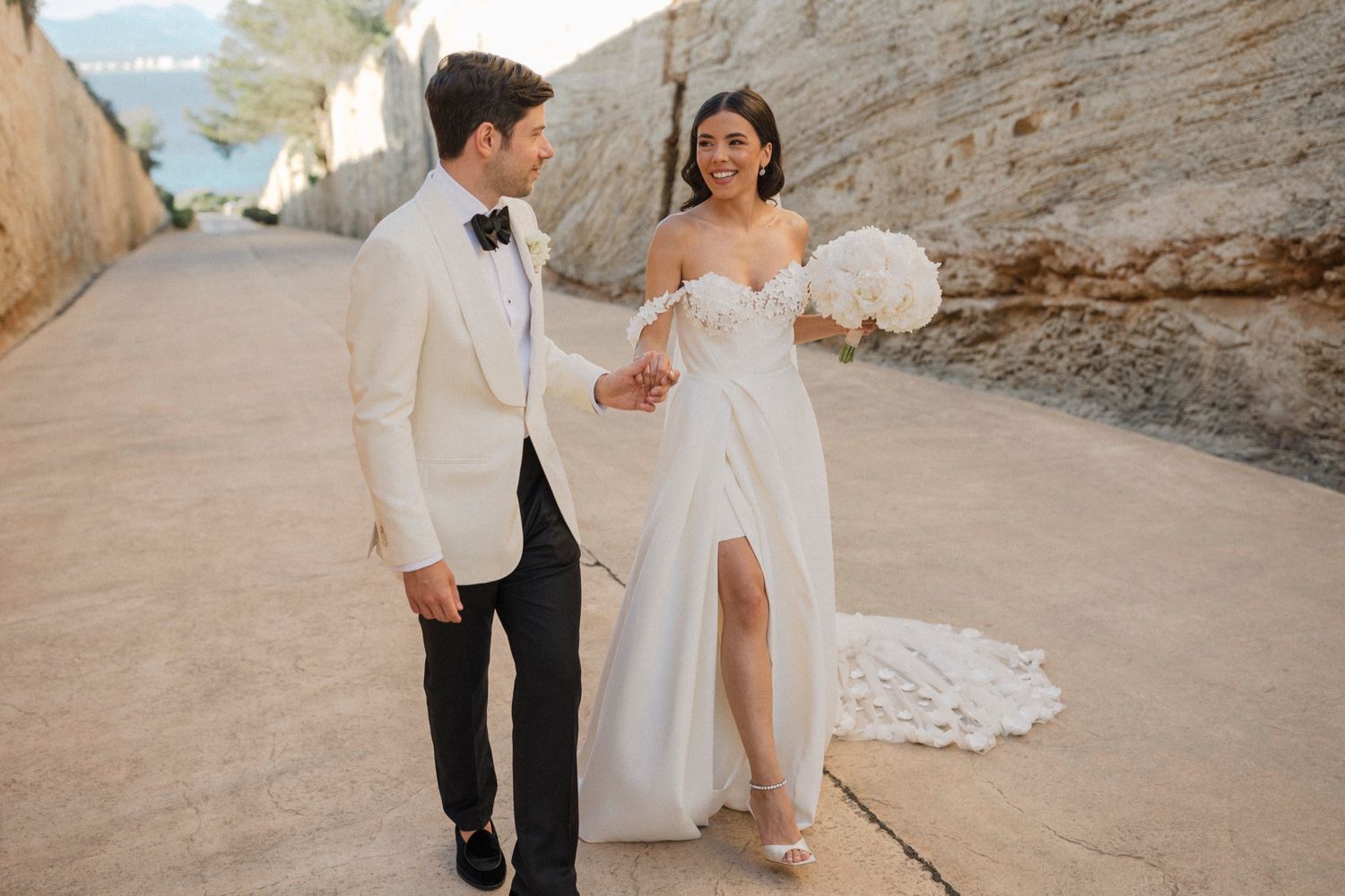 A bride and groom walk hand in hand in an elegant outdoor setting, she holds a white bouquet wearing a slit gown.
