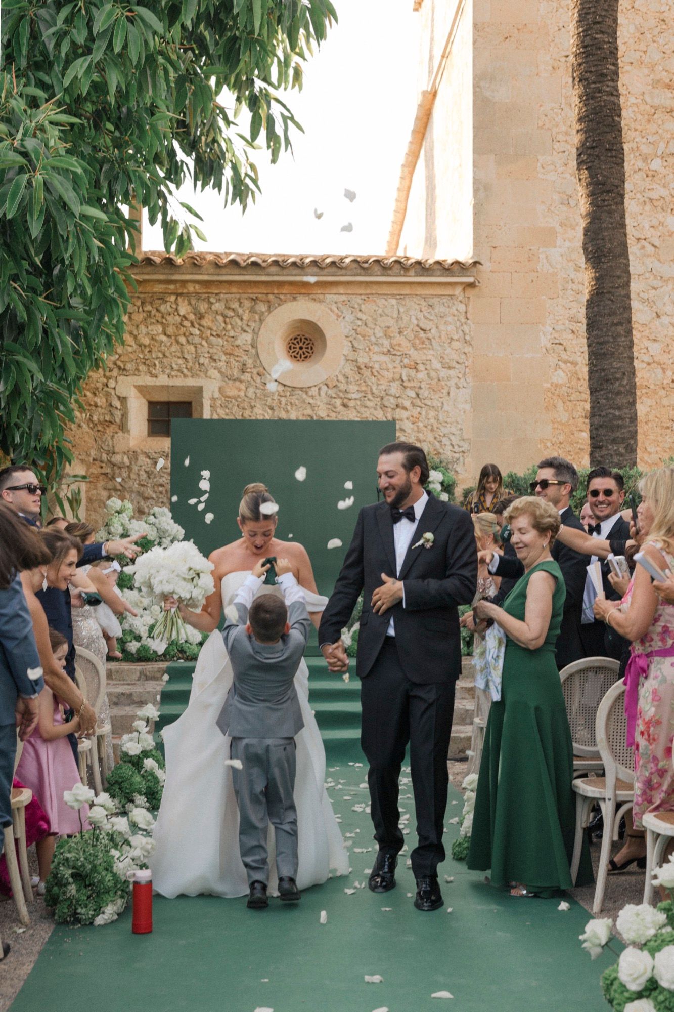 Wedding guests gather outdoors at venue with stone walls and palm trees.