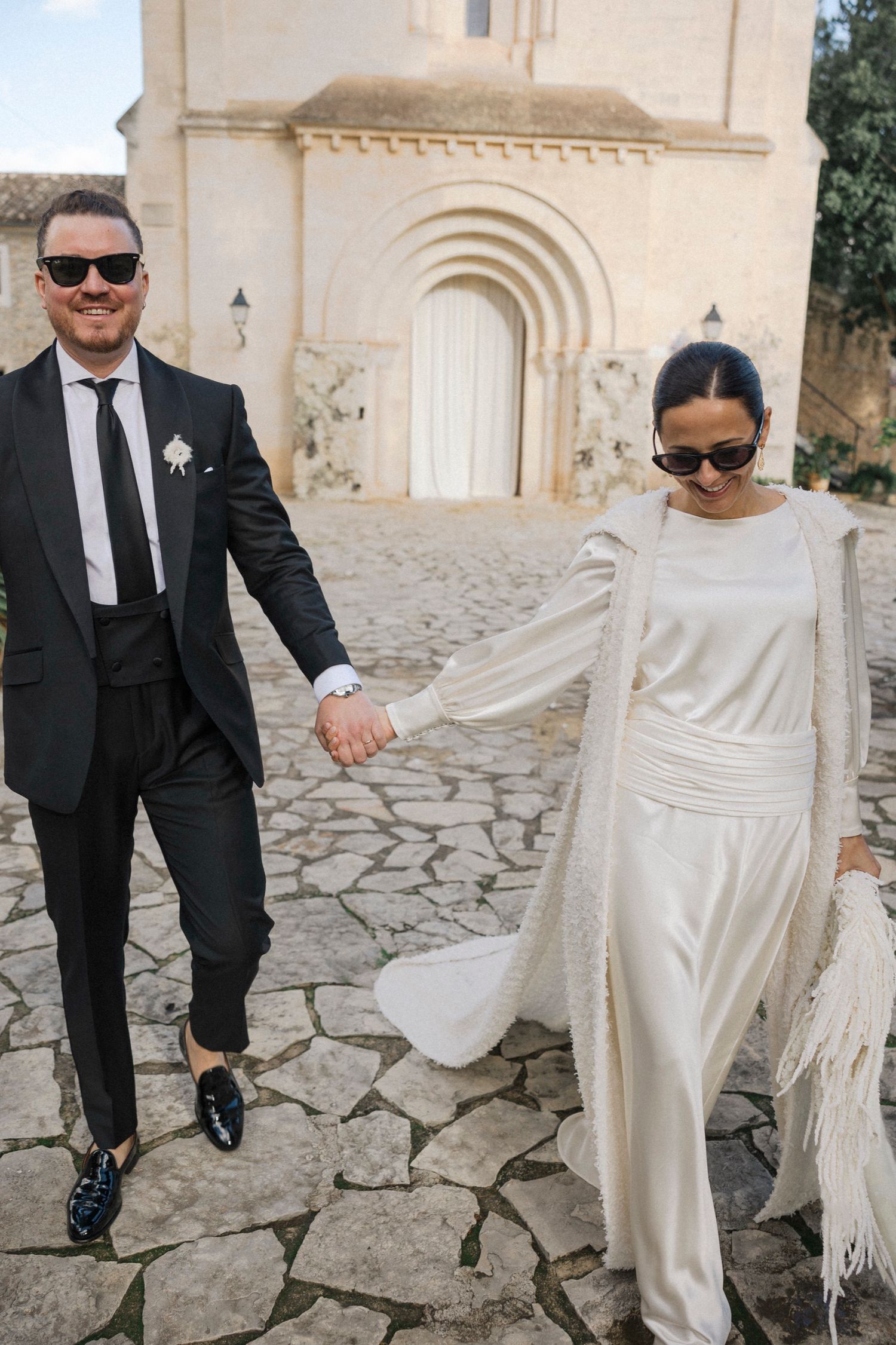 Stylish wedding couple walking hand in hand on cobblestone path by historic architecture.