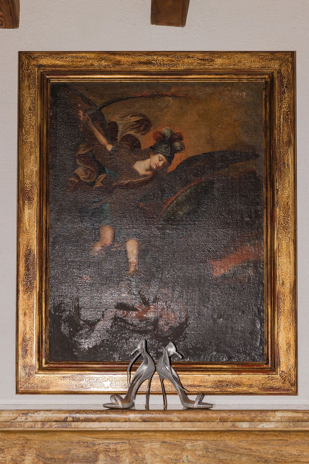 Silver wedding shoes displayed on mantel beneath gold-framed classical painting.
