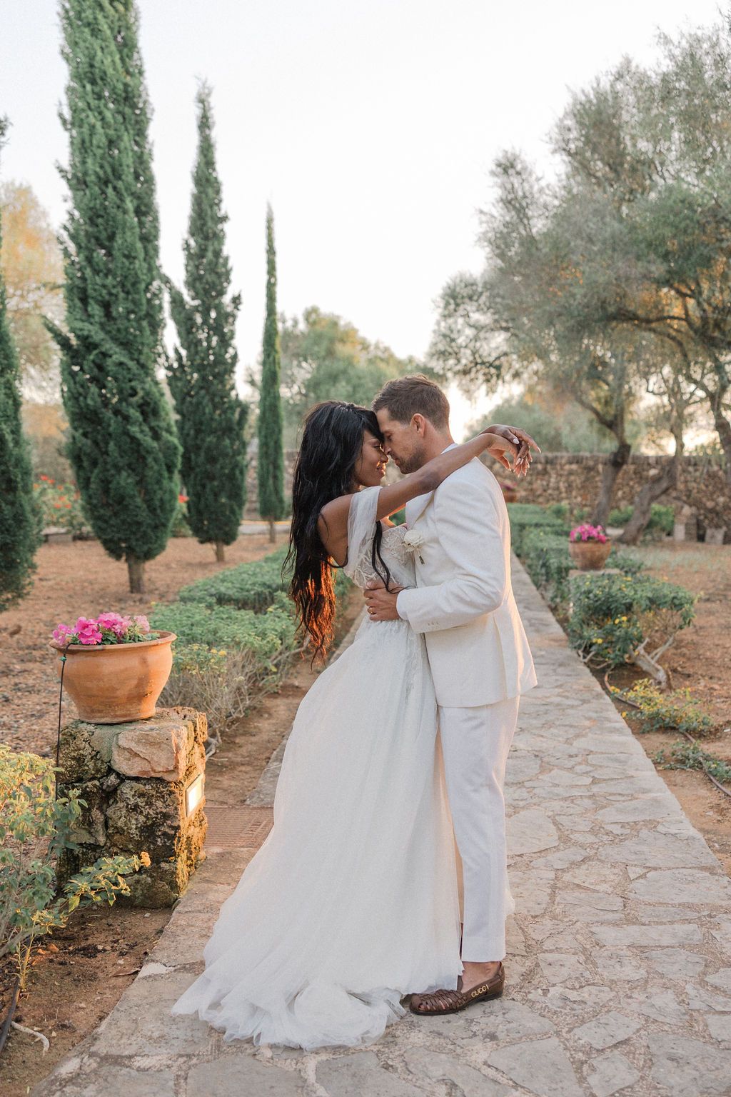Romantic couple in white sharing tender moments on stone pathway with cypress trees.