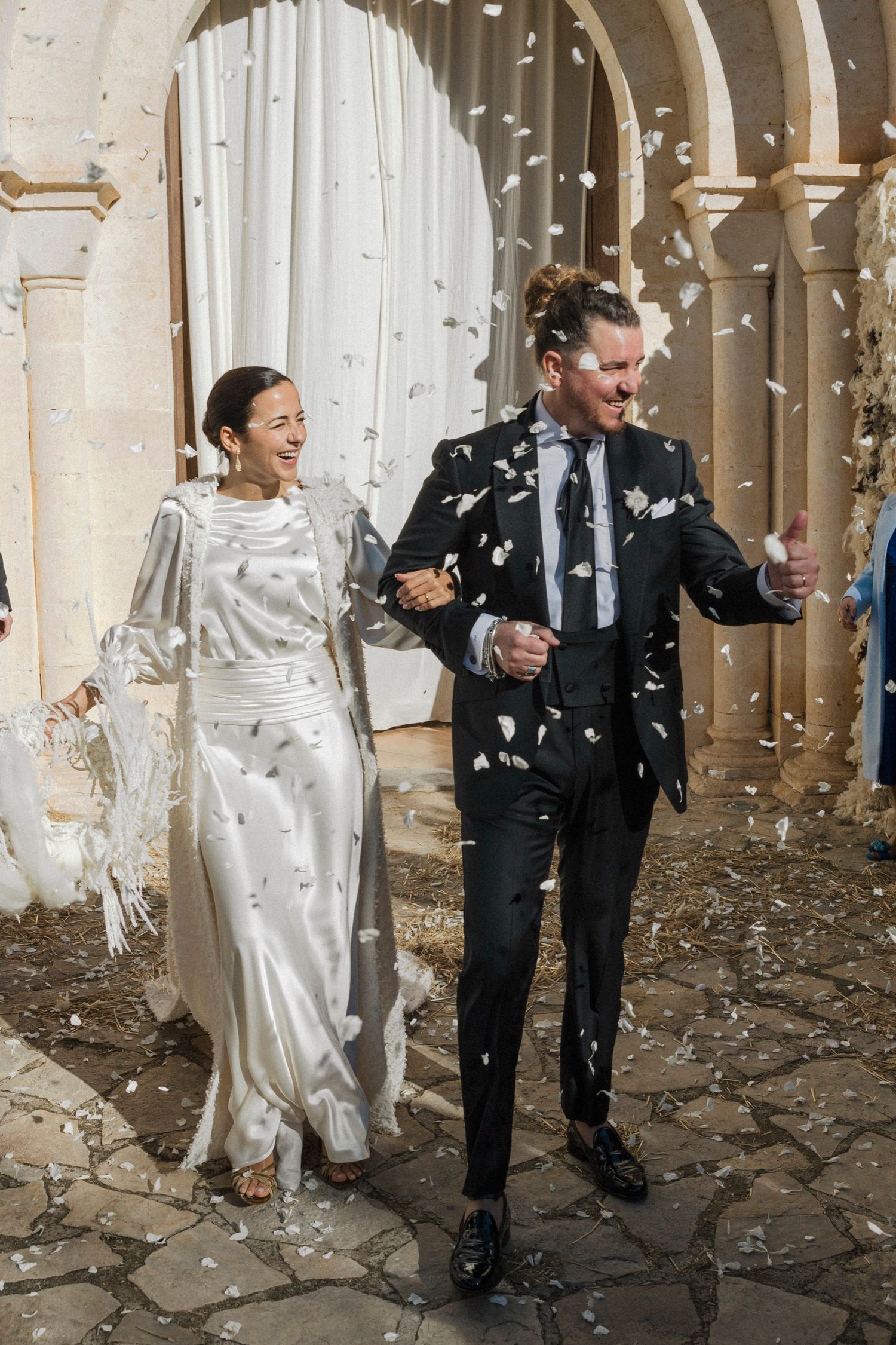 Newlyweds walking through shower of confetti under stone archway.