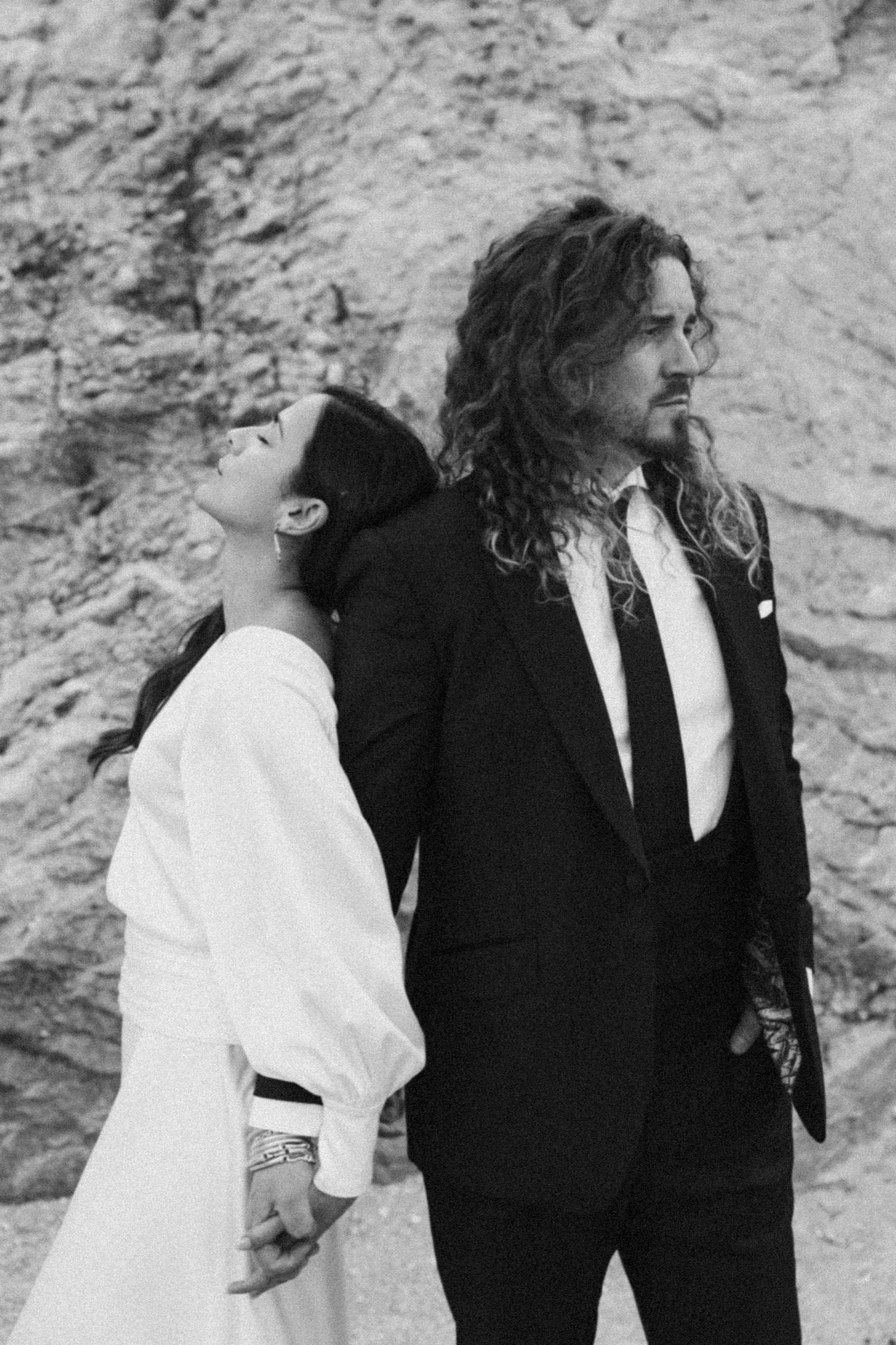 Artistic black and white portrait of couple in contrasting formal wear against rocky wall.