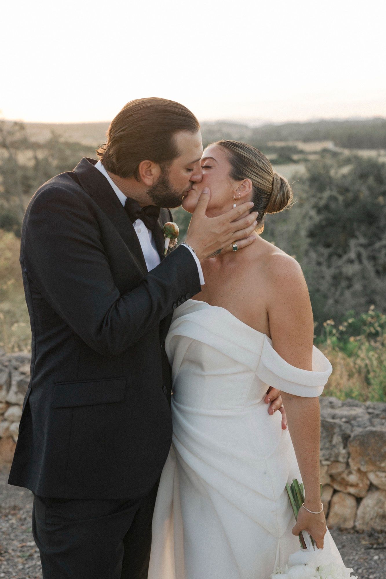 Romantic wedding couple embraces during sunset photoshoot with rustic stone wall and countryside backdrop.