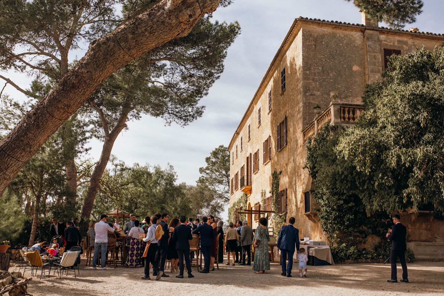 People gather outside rustic stone buildings along a tree-lined path at a Mediterranean estate property.
