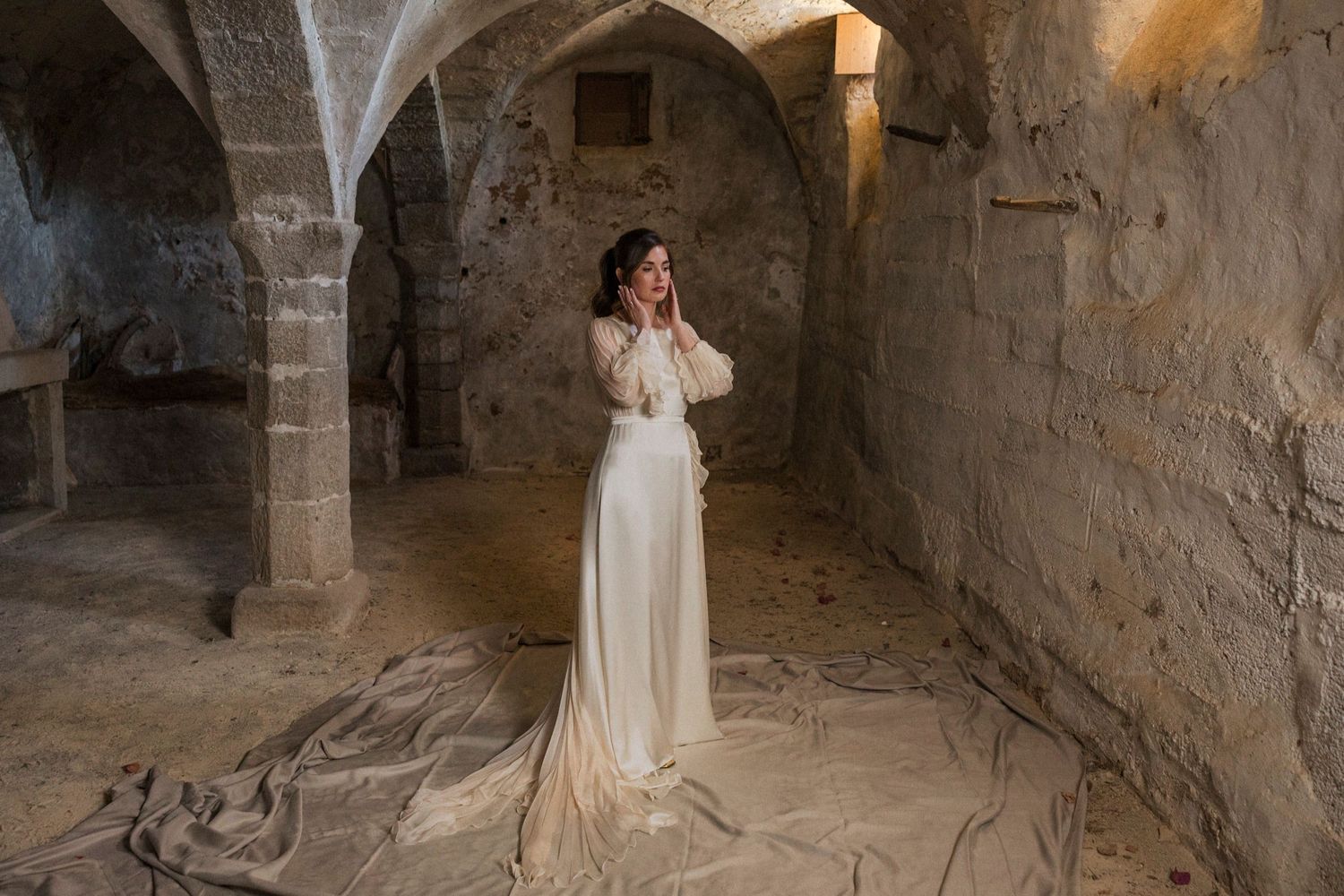 A person in a flowing white wedding dress stands in an ancient stone cellar with arched ceilings and dramatic lighting.