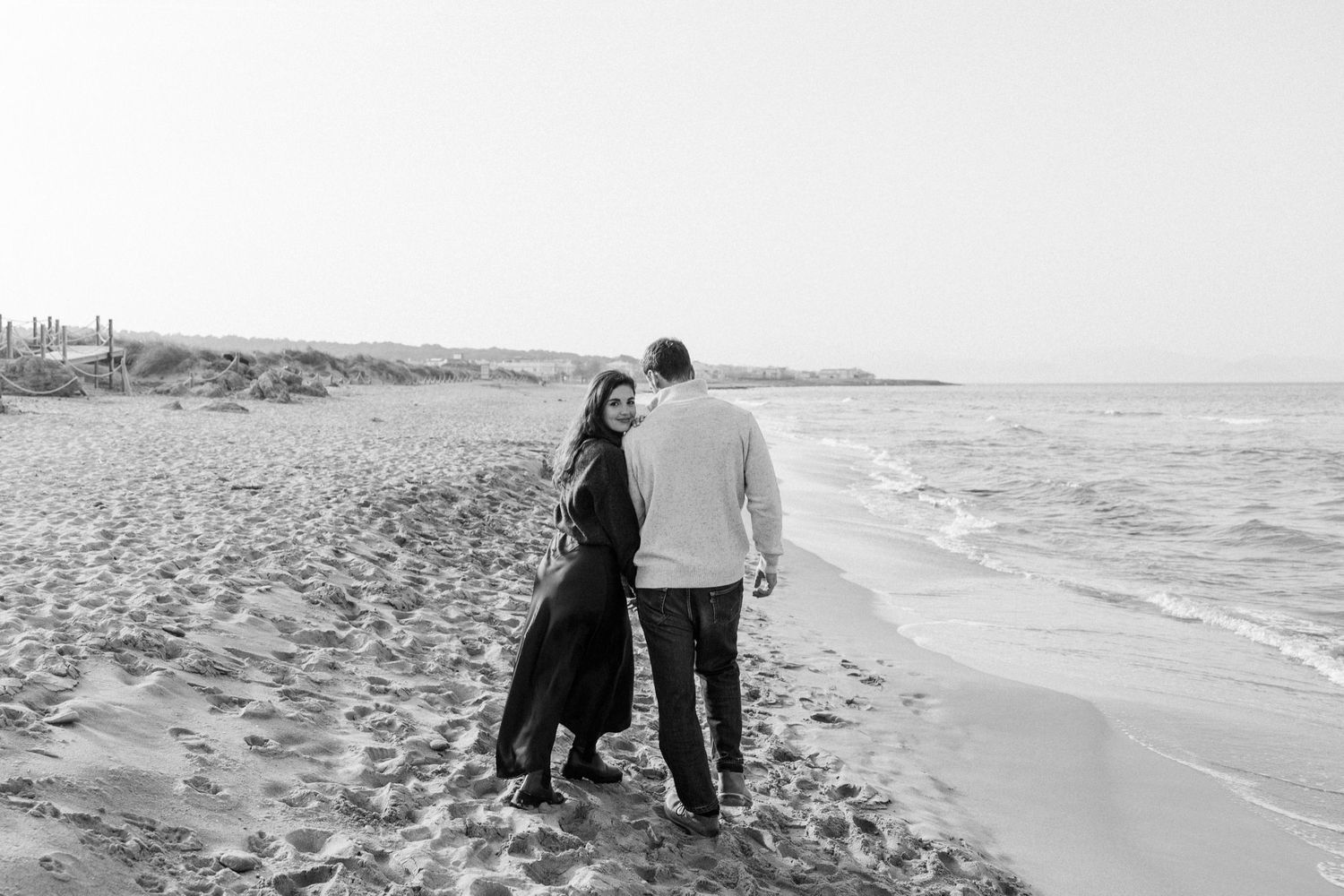A couple stands together on a sandy beach looking out at the ocean waves in black and white.