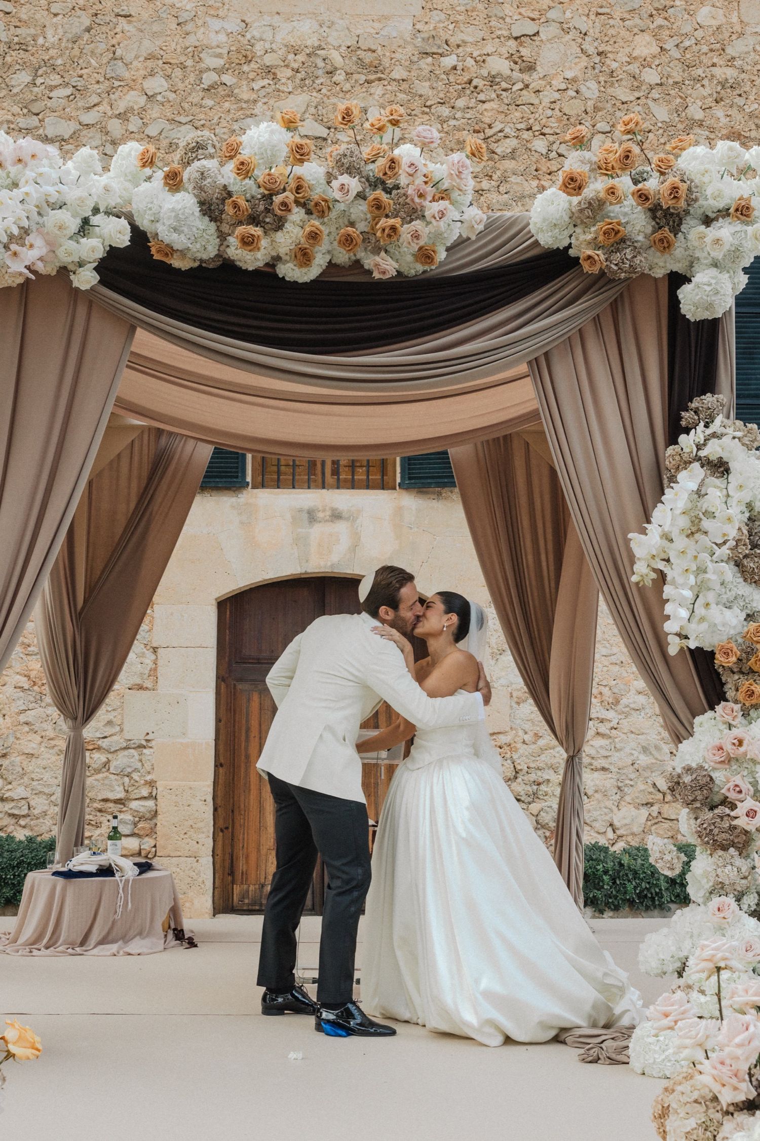 Romantic wedding couple shares intimate kiss under draped fabric gazebo decorated with white and peach floral arrangements.