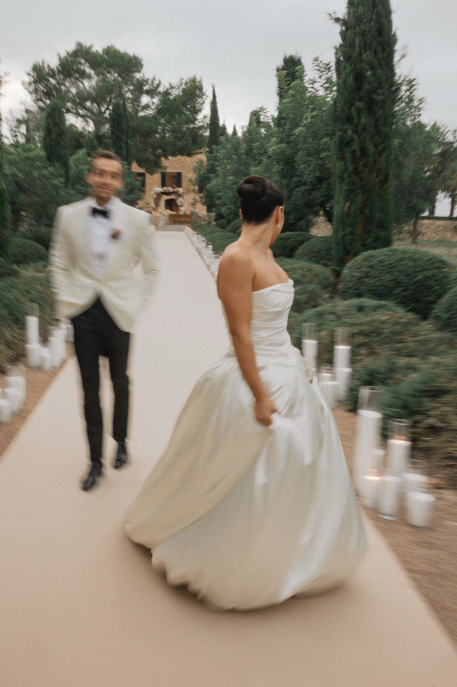 A couple in wedding attire twirls and dances together on a garden path surrounded by cypress trees.
