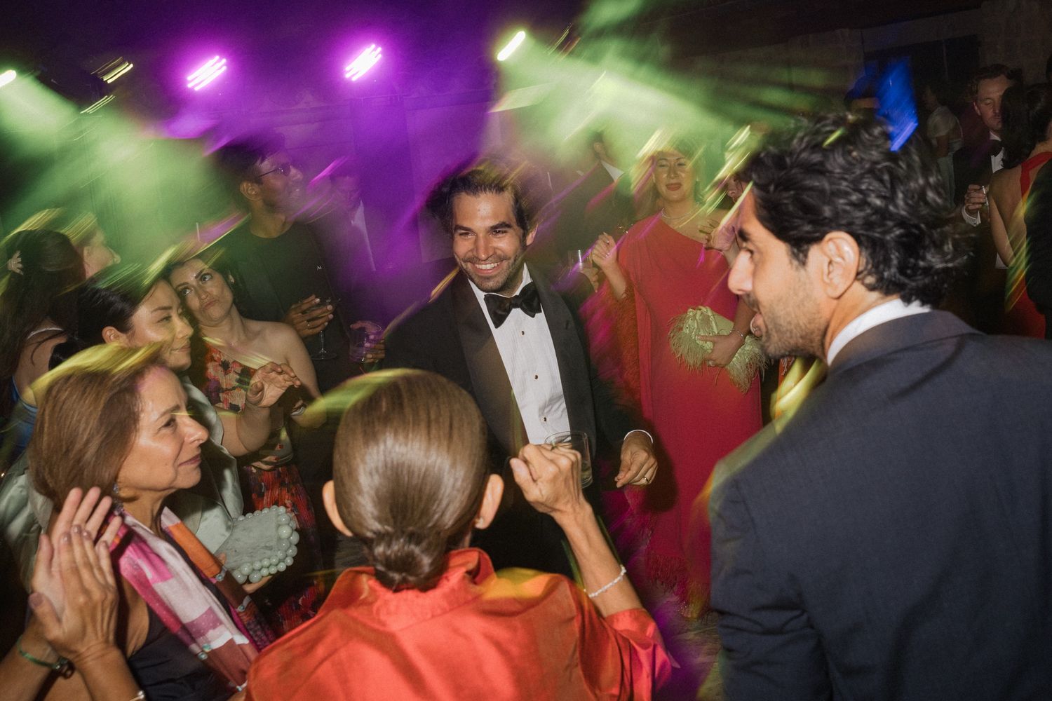People dancing and socializing at a nightclub with purple and green lighting effects in the background.