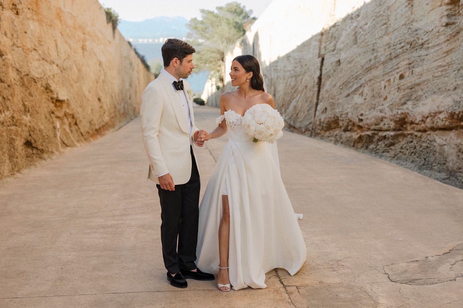 Bride and groom holding hands in a sunlit stone alleyway, bride in off-shoulder white gown, groom in white tuxedo jacket.