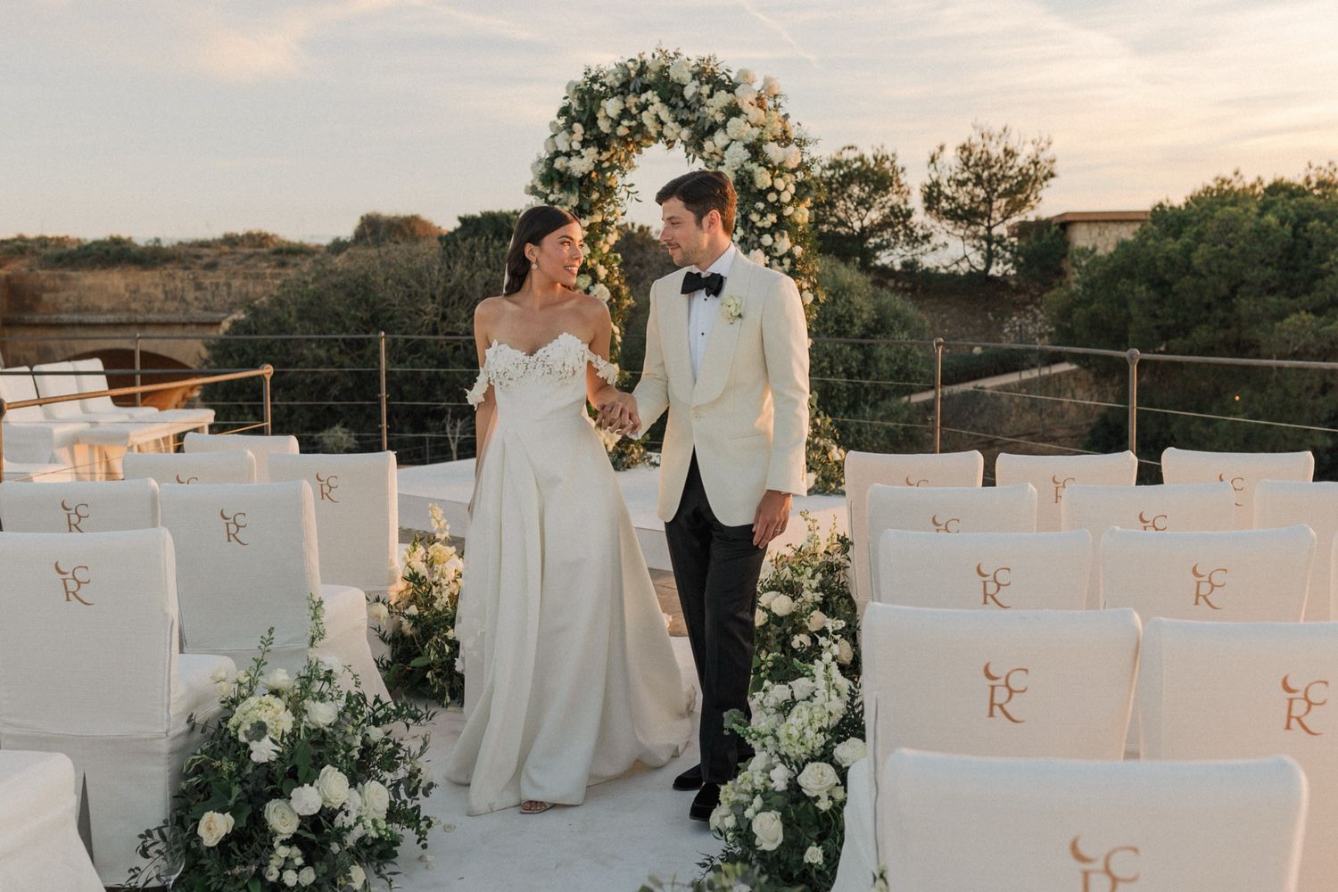 Bride and groom share a romantic moment at an elegant outdoor wedding ceremony with white chairs and floral arch.