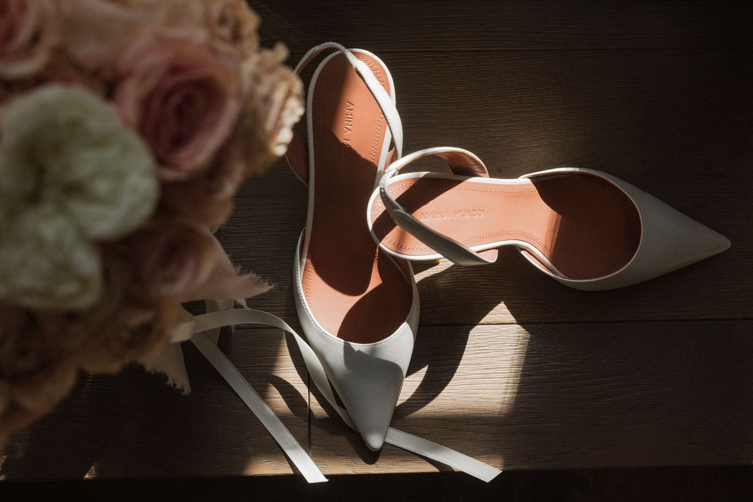 Wedding rings cast dramatic shadows on a textured surface beside soft pink roses in moody lighting.