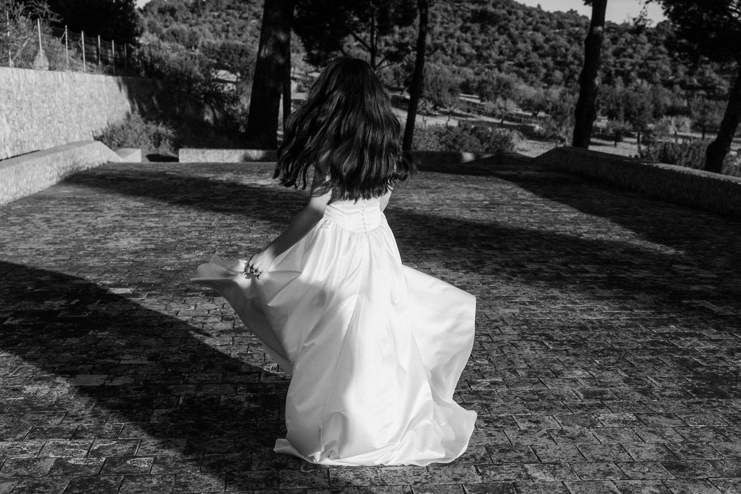 A bride in a flowing white dress twirls in an outdoor setting with trees and natural scenery in the background.