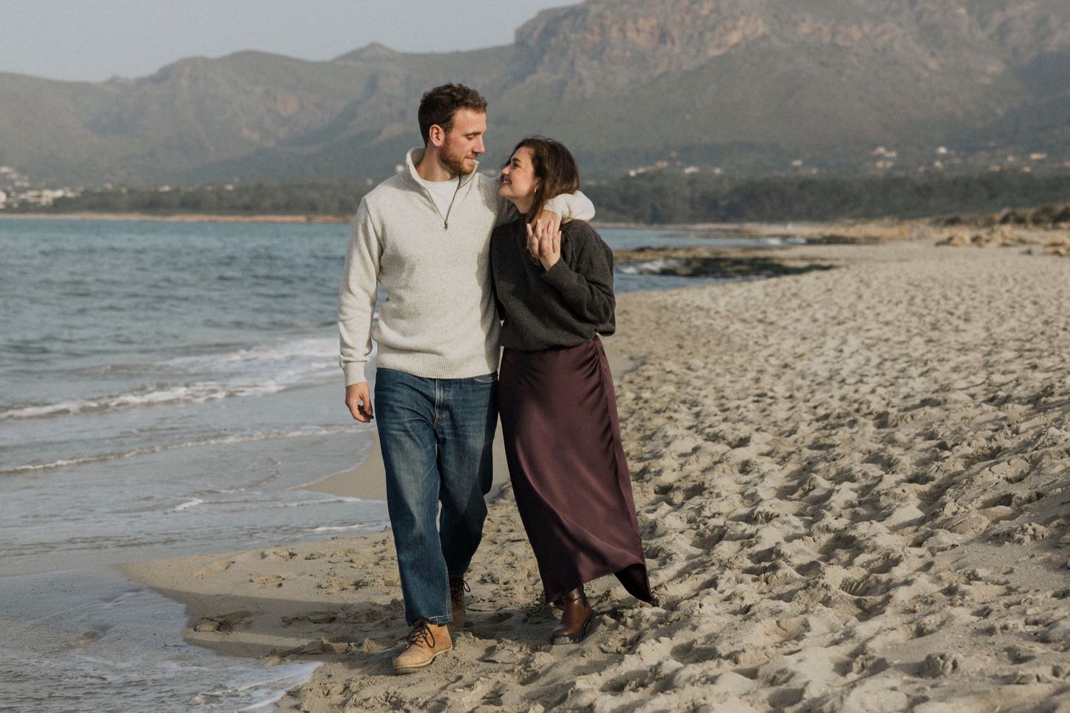 Couple walking and embracing on a scenic beach with mountains in the background during golden hour.