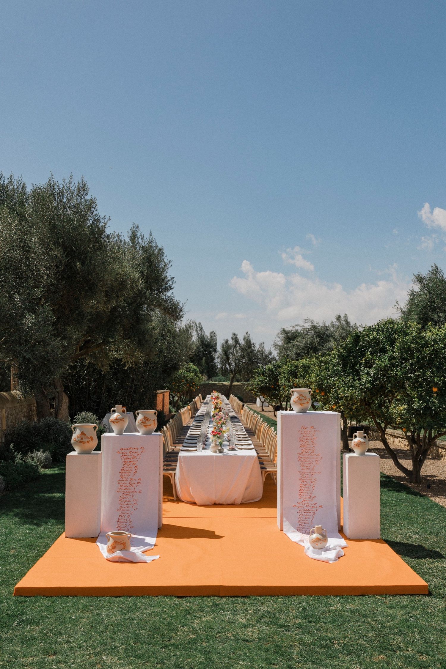 White ceramic vases arranged in a line on an orange platform against a backdrop of olive trees and blue sky.