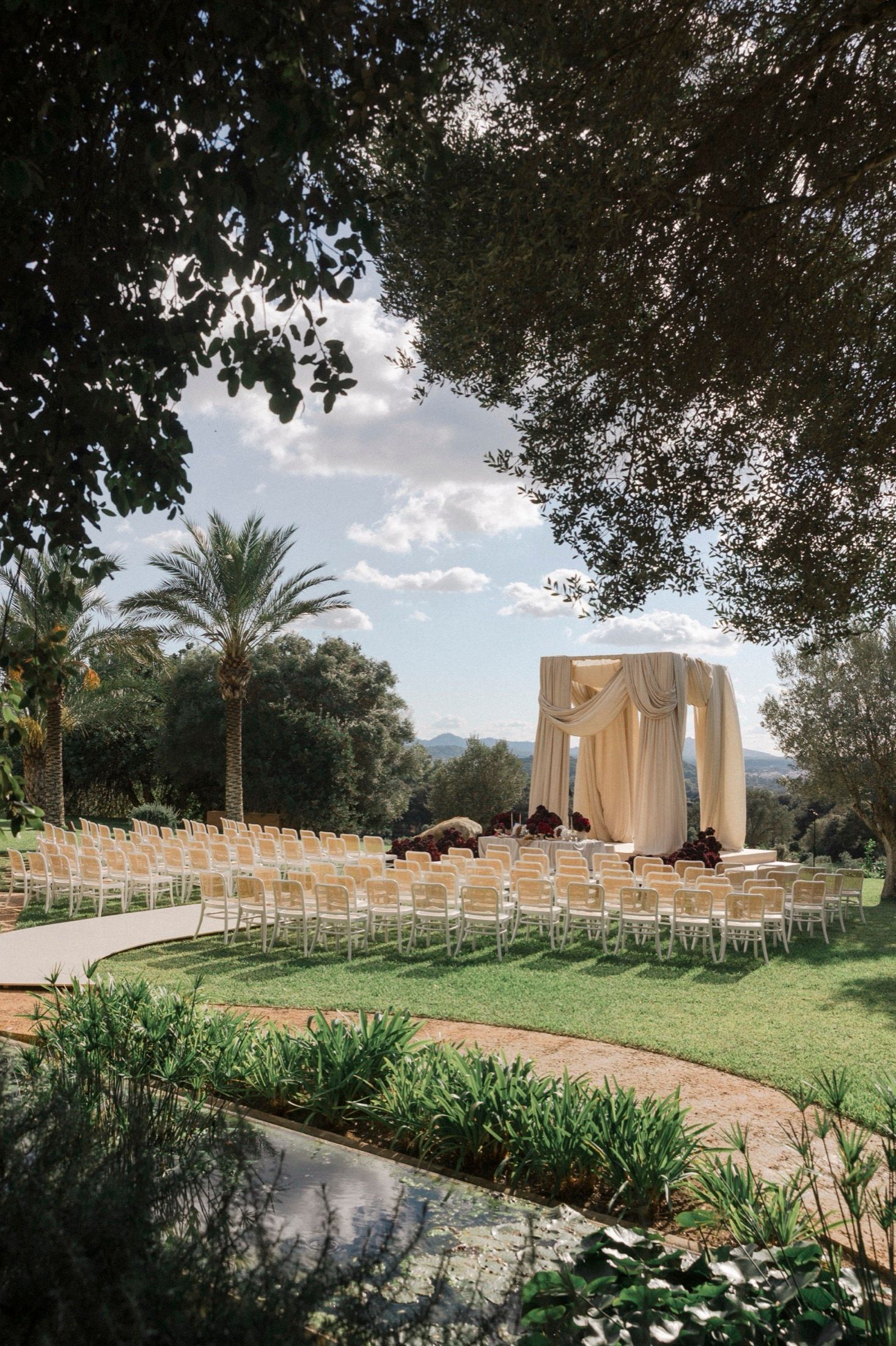 Outdoor wedding ceremony setup with white chairs and draped arch altar surrounded by palm trees and lush gardens.