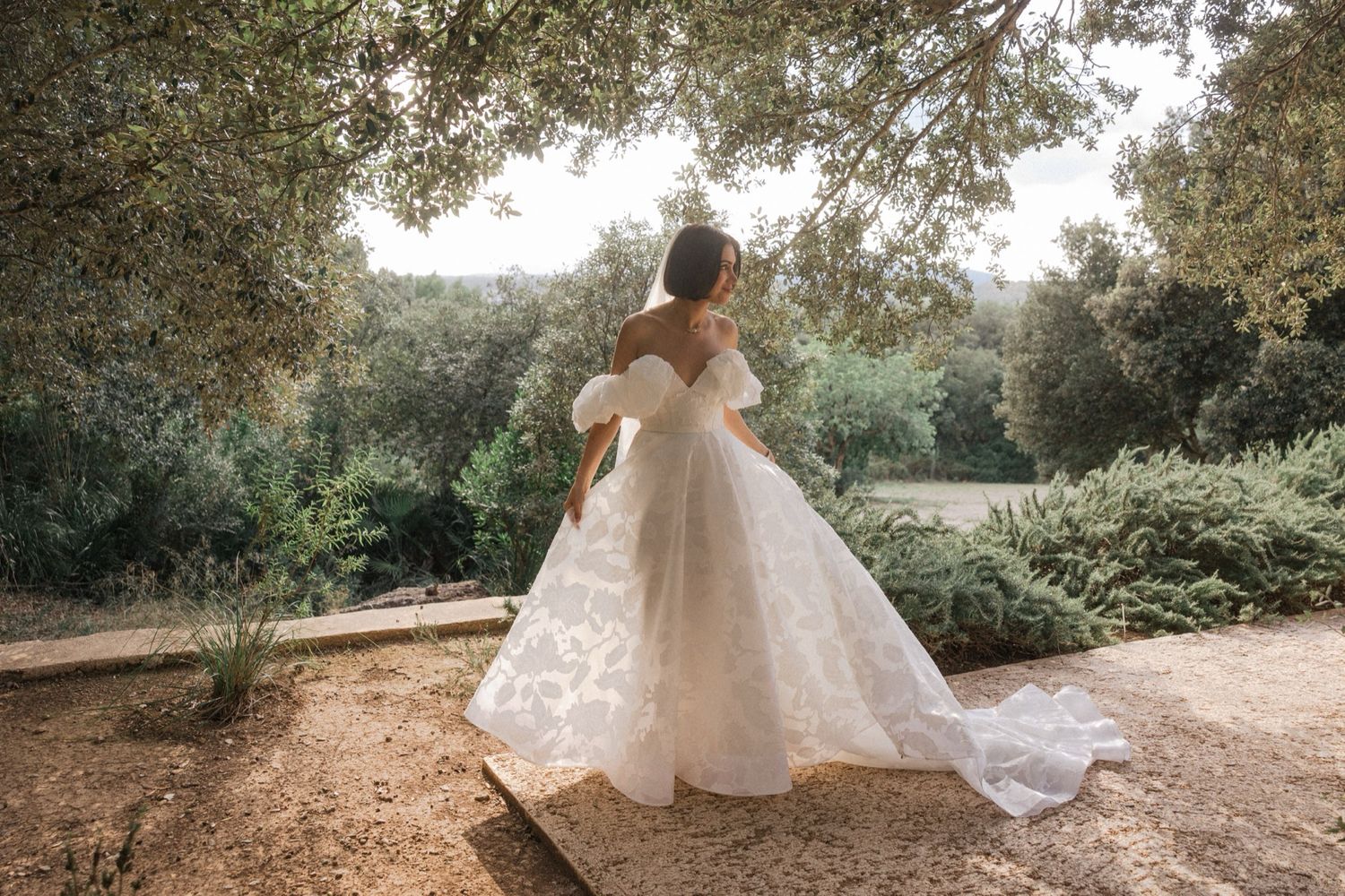 A romantic wedding photo series showing a bride in a flowing white gown walking through a sunlit garden path.