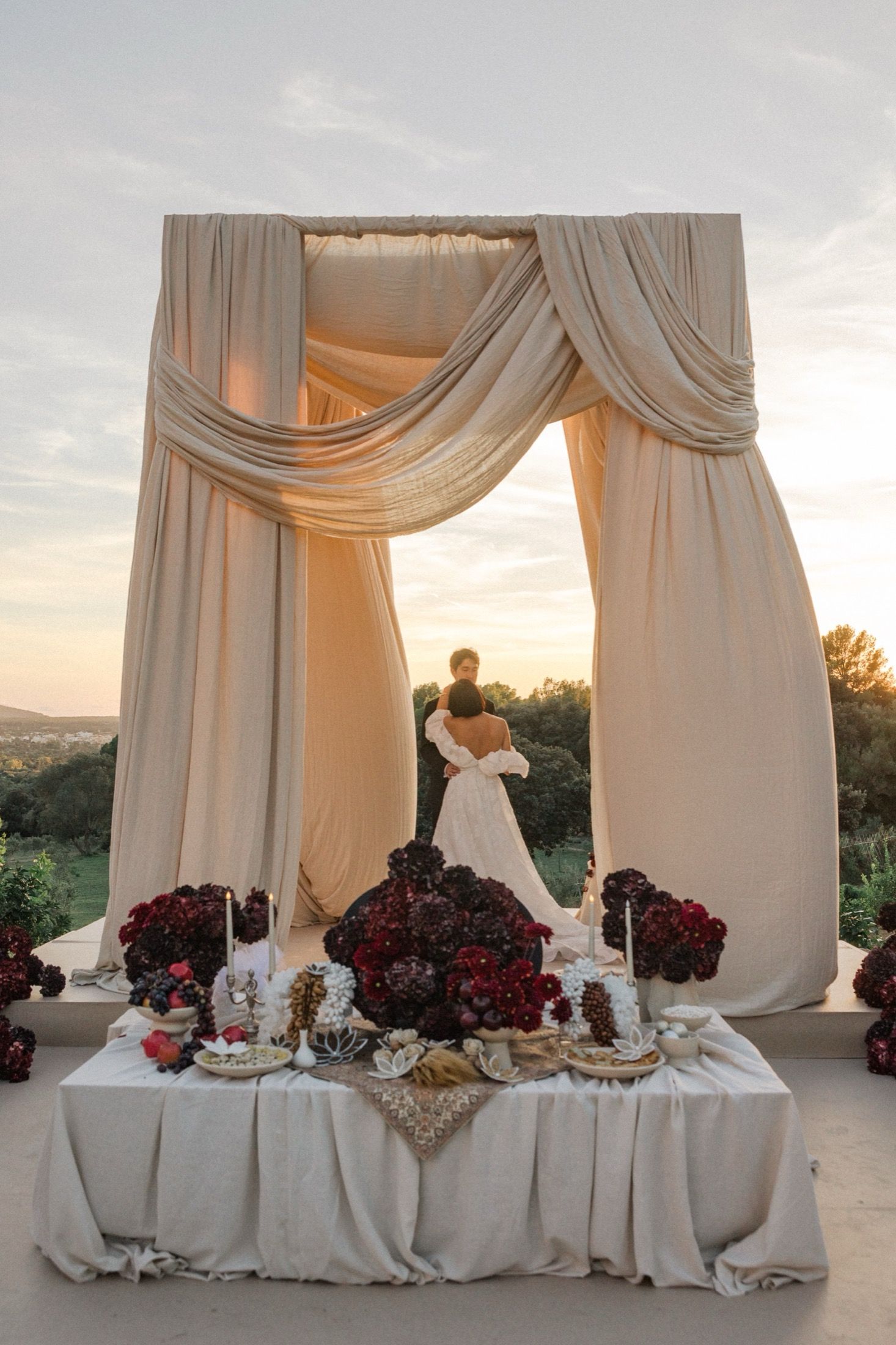 Elegant wedding reception table with white drapes and red floral arrangements against a sunset backdrop.