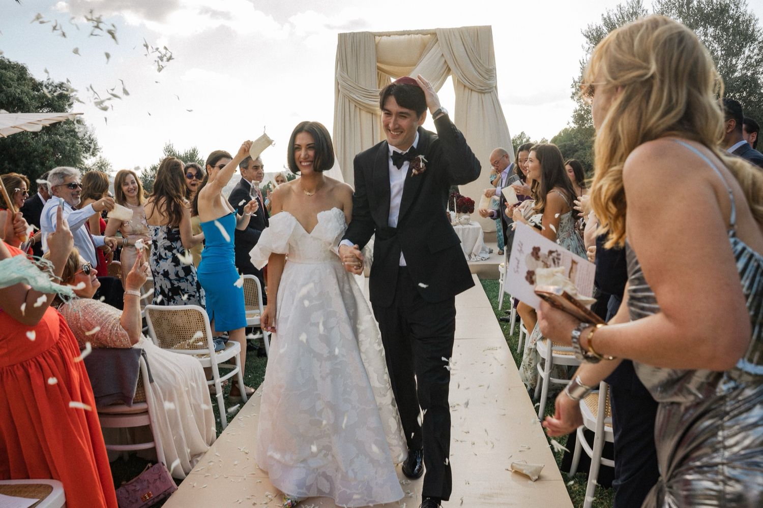 Newlyweds walk down the aisle at an outdoor wedding ceremony while guests shower them with confetti.